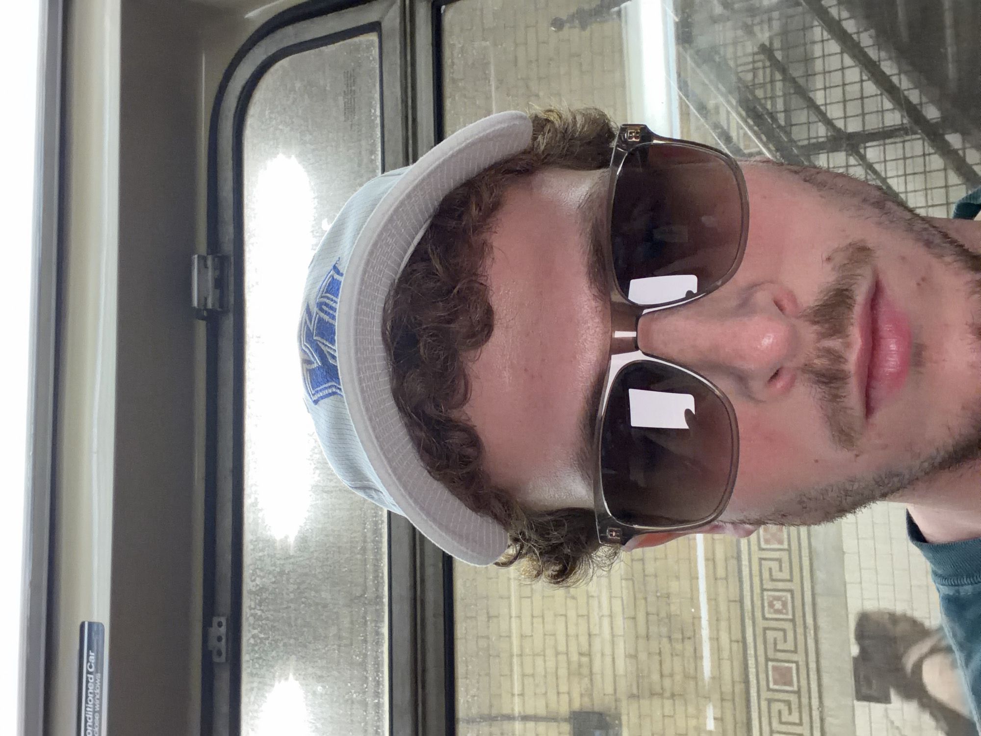 A person with curly brown hair, wearing sunglasses and a light-colored bucket hat, is positioned in front of a subway train window. Outside the window, the tiled wall of a New York City subway station platform is visible. The train car has interior elements and signage reading "Air Conditioned Car." The person is looking toward the camera.