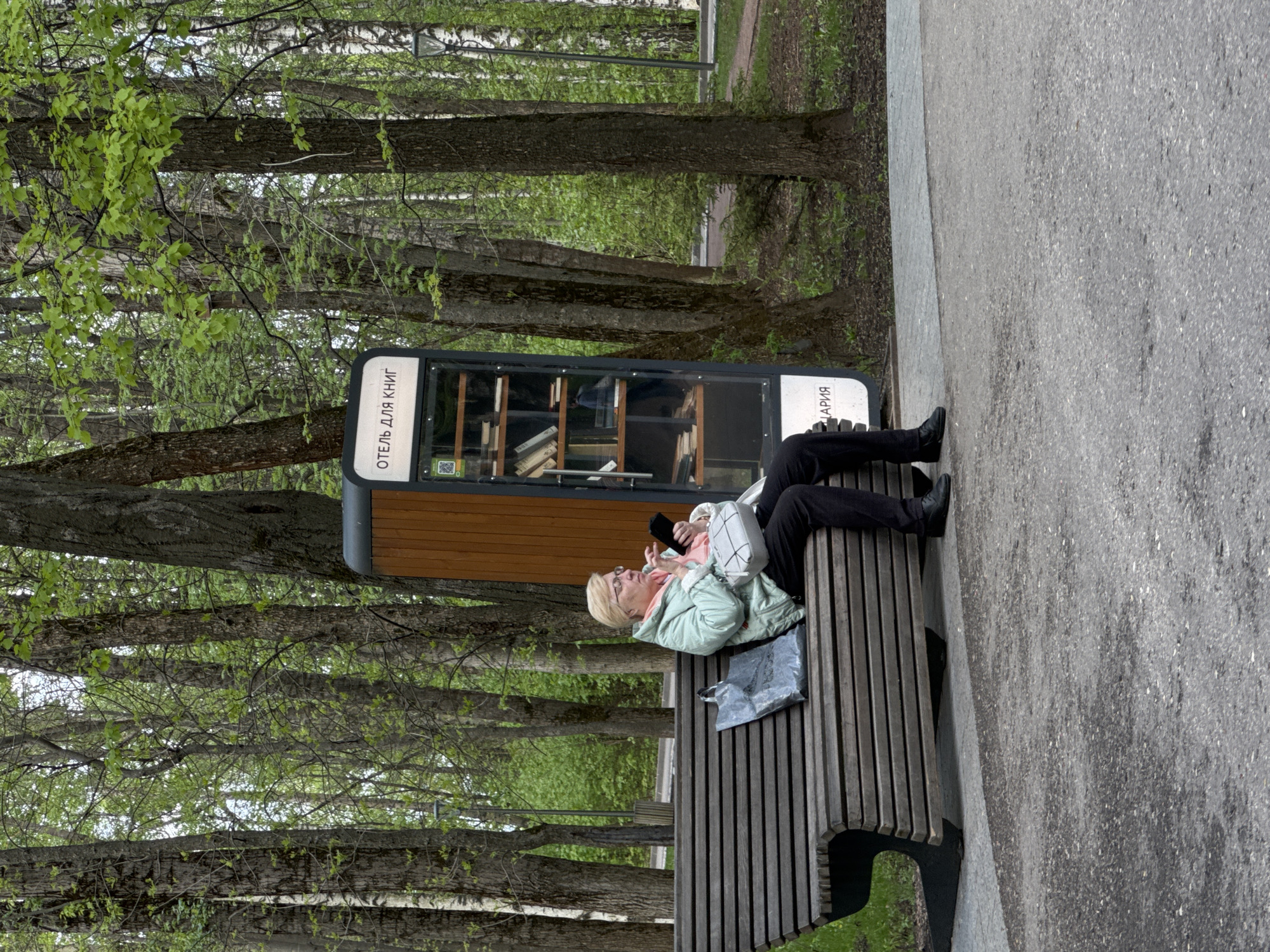 An elderly woman is sitting on a wooden bench in Nizhny Novgorod, Russia. She is wearing a light green jacket and black pants, and is holding a mobile phone in her hands. A book exchange box, labeled "Отель для книг" (Hotel for Books) is adjacent to the bench. Trees are visible in the background.