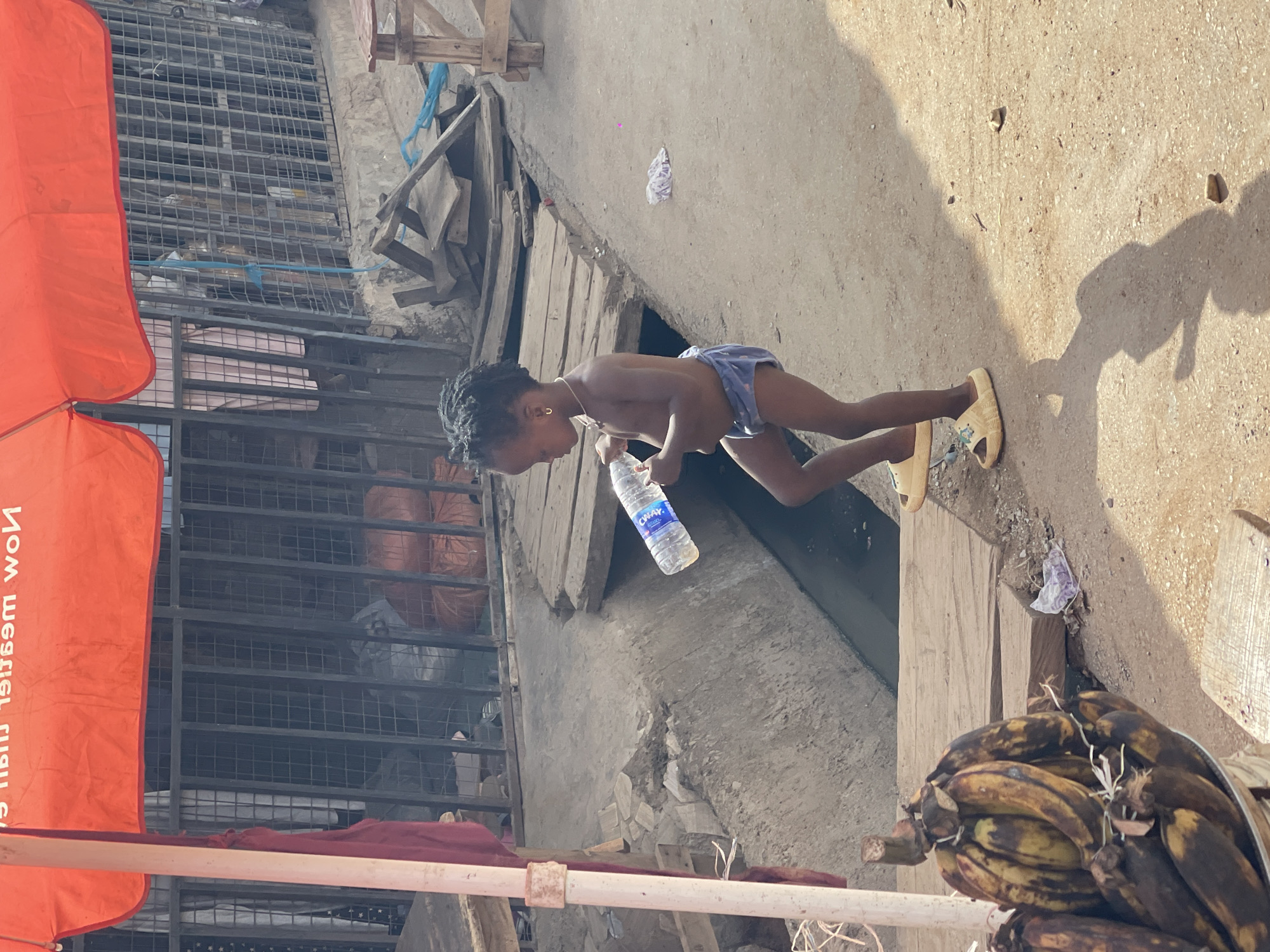 The image captures a sunlit outdoor scene, likely a street or market area in Kuchingoro, Nigeria. The time of day is bright, indicated by strong shadows cast on the ground.
The central subject is a young child, possibly a toddler or preschooler, with dark skin and braided hair. The child is shirtless, wearing light blue shorts or a diaper, and light yellow sandals. They have a necklace and small hoop earrings. The child is holding a clear plastic water bottle with a blue label that reads "CHAY." The child's posture suggests they are either walking or standing, focused on the bottle.
In the foreground, to the right, a large bunch of bananas, some ripe and some bruised, are visible, tied with string. To the left, an orange umbrella or canopy stands partially visible, displaying reversed and partially legible text that appears to be an advertisement, beginning with "Now m..." and ending with "wow" (when read in correct orientation).
The background features various structures, including metal grates and fencing, possibly part of stalls or buildings. Wooden planks and debris are also scattered. The ground is a light-colored, possibly concrete or dirt surface, with small pieces of litter. A distinct shadow of the child is cast on the ground to the right.