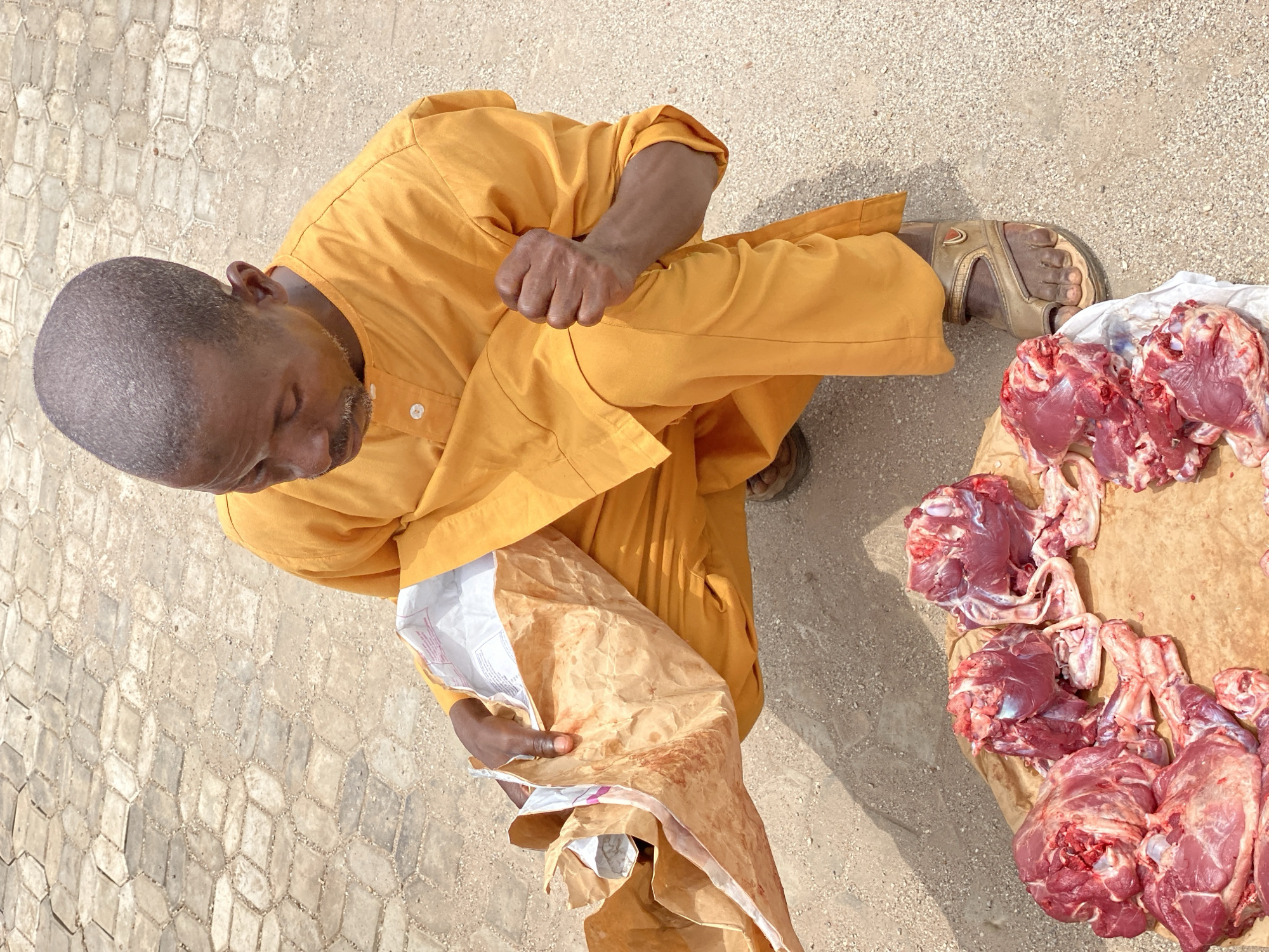 An elevated, downward-facing perspective of an outdoor scene in Maiburiji, Nigeria, captures a man interacting with raw meat. The man, who is bald, wears a yellow tunic and brown sandals. He stands on a light-colored, patterned paved surface and holds a partially open, blood-stained paper bag. To his right, a selection of raw red meat pieces rests on a brown surface, likely a piece of butcher paper or a cutting board. The environment consists of textured paving stones or tiles. There is no explicit action occurring beyond the man holding the bag and the presence of the meat. No interactions are readily apparent beyond the man and the meat.