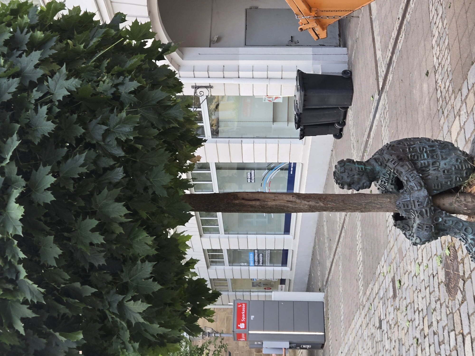 This media file depicts an outdoor scene in Ansbach, Germany. The main subjects are a bronze sculpture of a person intertwined with a tree, and a large tree with dark green leaves in the foreground. The sculpture appears to be made of many letters and words, adding a unique texture to the figure.
In the background, a modern building with white facade and large windows is visible. Some windows have signage, and one clearly displays "Sparkasse," a German savings bank. Other text visible on the signage includes "WIR GEBEN RÜCKENWIND" and "AUCH AUSSERHALE DER."
Several black trash bins with wheels are also present on the paved sidewalk. An orange object, possibly part of construction equipment, can be seen in the upper right corner. The ground is paved with cobblestones. The scene suggests an urban setting, possibly a street or public square, during daylight hours. The lighting is neutral, indicating it's likely daytime, though no strong shadows suggest a specific time. The overall atmosphere is neutral, focusing on the juxtaposition of nature (tree and sculpture) and urban infrastructure.