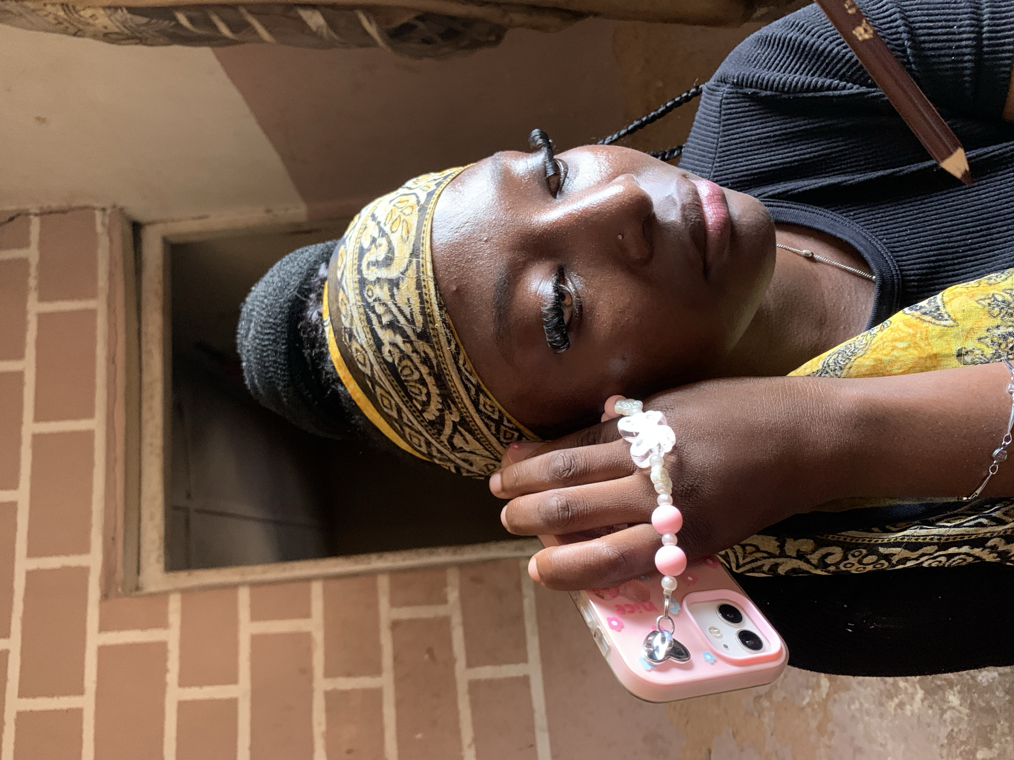 The image depicts a young Black woman indoors, holding a pink smartphone with a beaded charm bracelet attached to it. She is wearing a black ribbed shirt and a yellow and black patterned headscarf. Her hair is styled in a bun, with a braid visible down her back. Her makeup includes prominent false eyelashes and a nose piercing. She is holding a brown pencil in her right hand, near her chin. The background shows a brick wall and a doorway, suggesting a simple, possibly unfinished, interior. The lighting appears natural, casting shadows on her face. The scene is likely in Jos, Nigeria, based on the context provided.