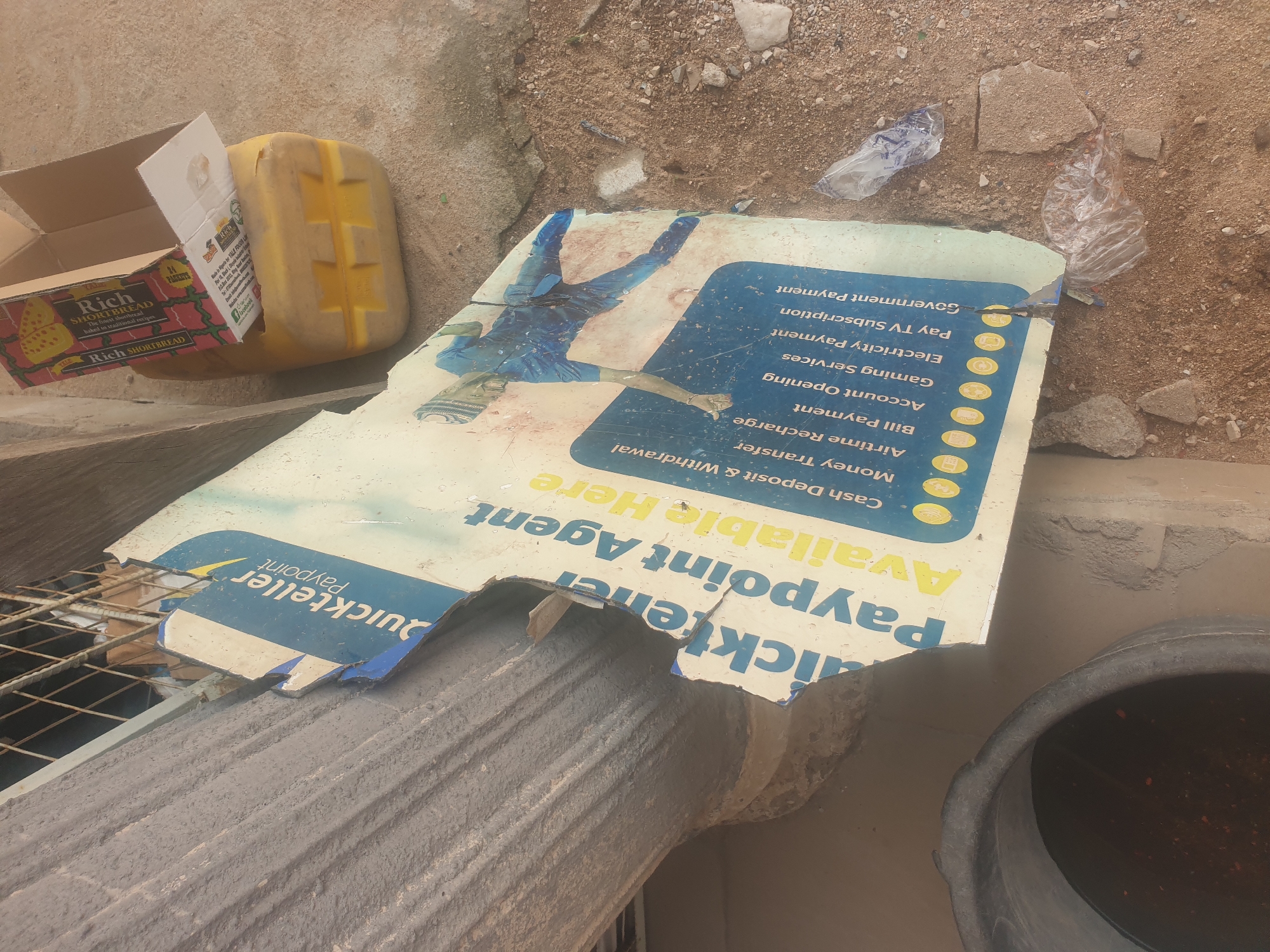 The image shows a damaged "Quickteller Paypoint" sign lying on the ground in Jauro Yinu, Nigeria. The sign is broken and peeling, with some sections missing. Text on the sign advertises services such as cash deposit and withdrawal, money transfer, airtime recharge, bill payment, account opening, gaming services, electricity payment, pay TV subscription, and government payment. A partially opened cardboard box containing what appears to be a box of Rich shortbread biscuits is placed on a yellow plastic container in the background. Debris, including stones and plastic bags, is scattered on the sandy ground around the sign. A large black bucket is visible in the lower left corner.