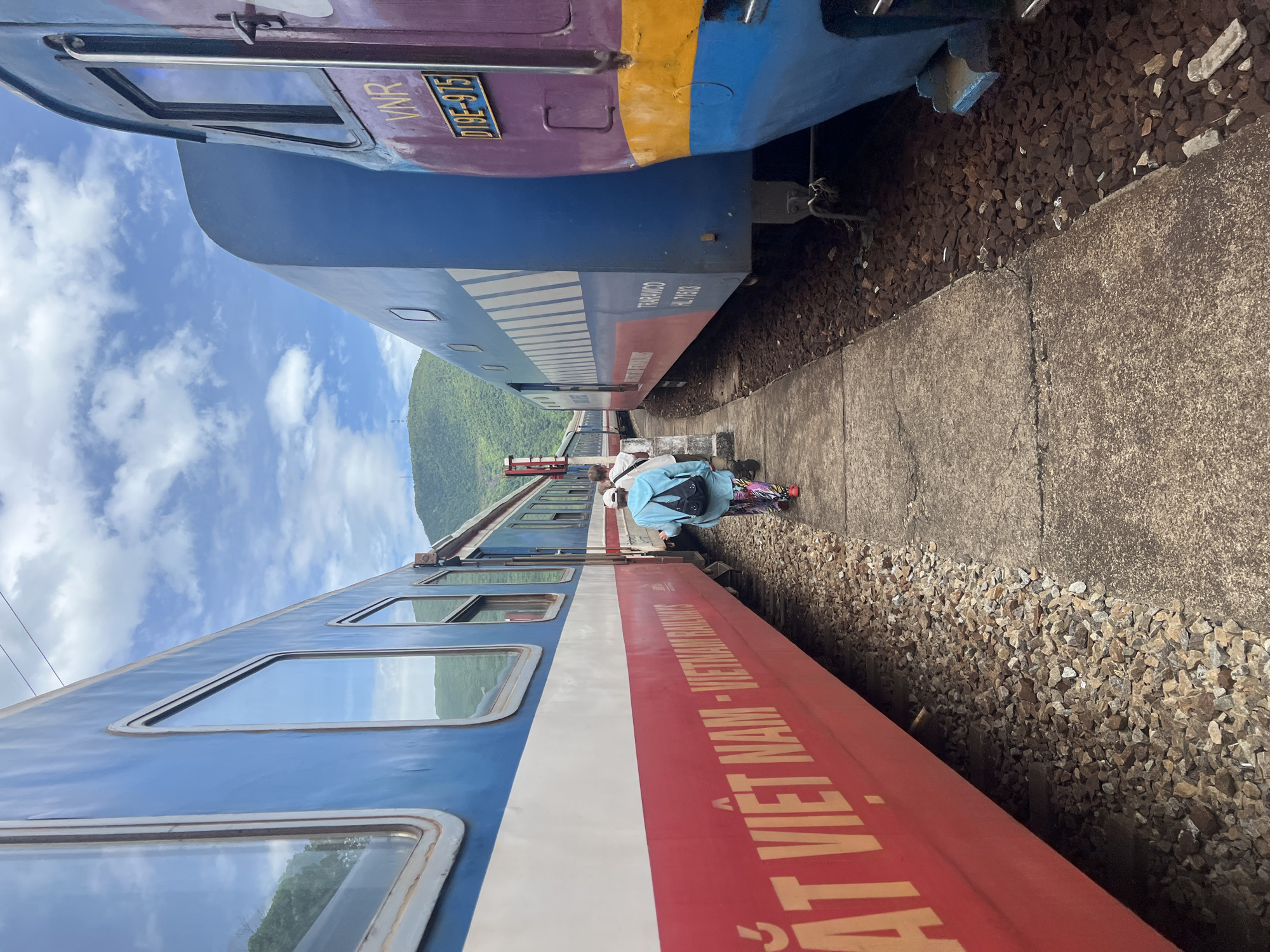 Two colorful trains, one primarily red and white with "VIET NAM RAILWAYS" text, and another blue and purple with "VNR" markings, are parked side-by-side on train tracks. A narrow concrete pathway separates the trains. A person wearing a light blue top and patterned pants is walking away from the camera on the pathway, heading towards the trains, with another person visible behind them. In the background, lush green mountains rise under a cloudy blue sky. This scene is likely at the Huế Railway Station, a significant transportation hub in Huế, Vietnam, known for its picturesque setting amidst hills and its historic significance.