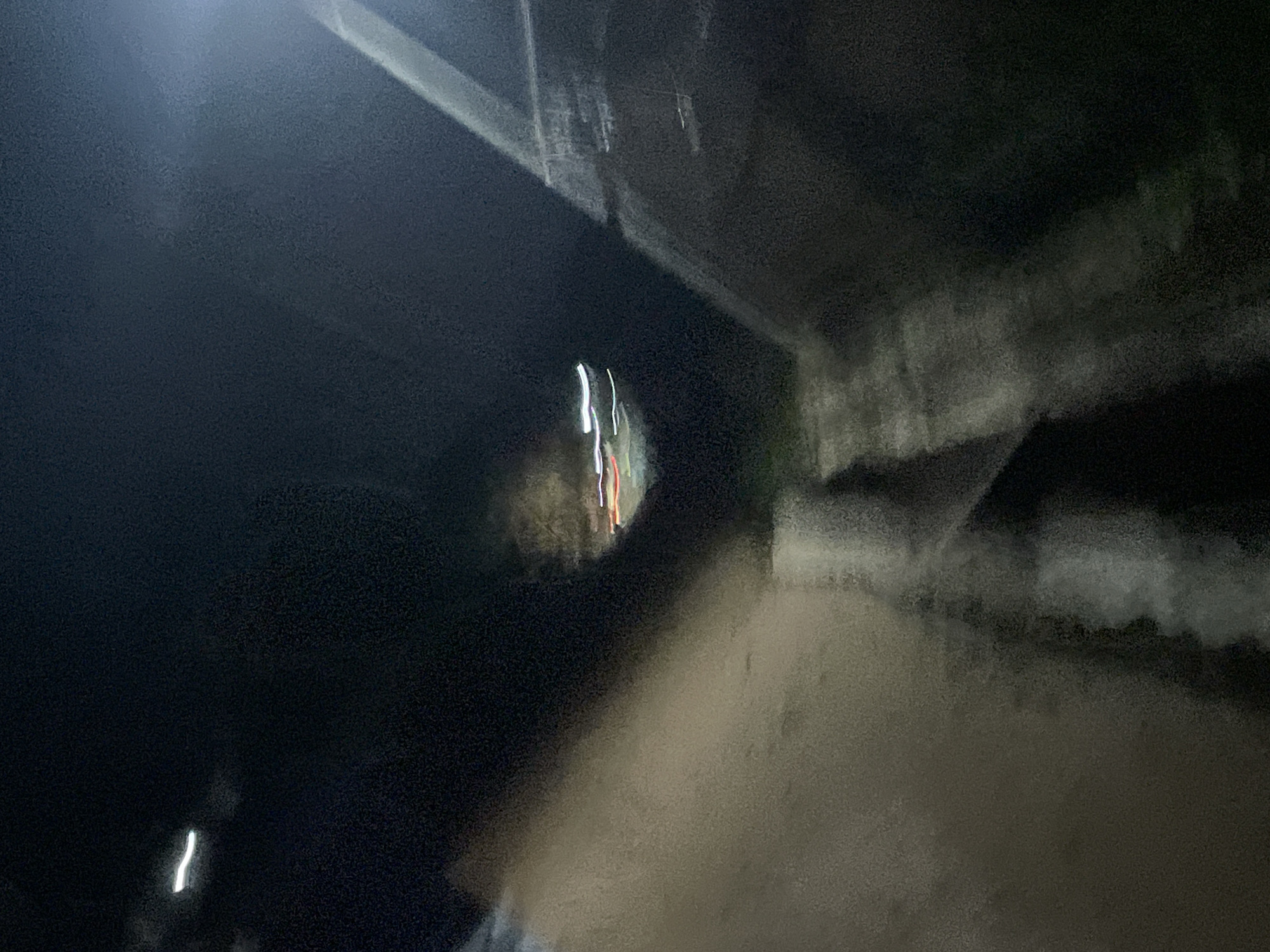 The image depicts a blurry, nighttime scene that appears to be a pathway or road in an urban setting, possibly Lokoja, Nigeria. The lighting is dim and suggests artificial illumination, with streaks of light indicating movement, likely from vehicles. There are indistinct shapes on either side of the pathway, which could be buildings, walls, or natural elements. The overall impression is one of low visibility and possibly motion blur due to the low light conditions and camera movement. The scene is dark and somewhat abstract, with limited discernible details.