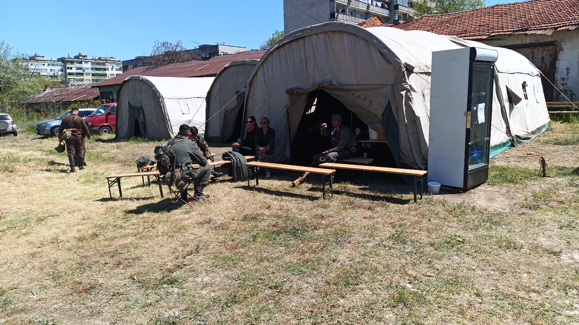 The image depicts an outdoor scene, likely during daylight, near the city of Yambol, Bulgaria. Several light-colored, military-style tents are visible, one of which has a refrigerator positioned at the entrance. Several individuals are present, including two women seated in front of a tent. Adjacent to them and inside the tent, there are people sitting on benches, possibly engaged in conversation. One man is walking toward the tents while carrying a helmet, and several other men are sitting in the shadows near the tents. In the background, there are cars, buildings, and trees. The ground is covered with dry grass and some green patches.