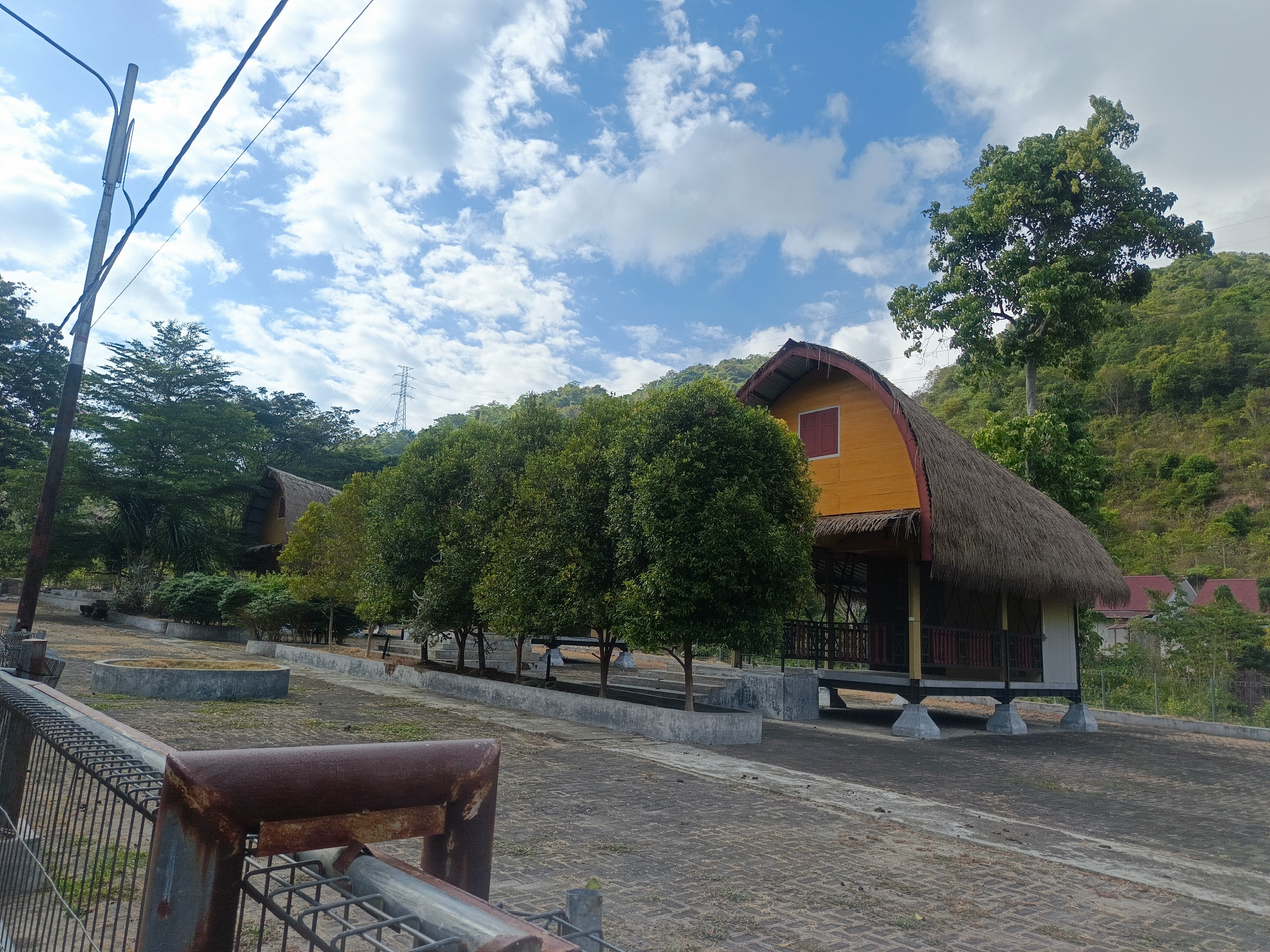 The image depicts a clear blue sky with scattered white clouds above a rural landscape in Gorontalo, Indonesia. In the foreground, a rusty metal fence runs across the lower portion of the frame. Behind the fence, a paved area with interlocking brick patterns leads towards several thatched-roof structures with yellow walls. These structures appear to be small, stilted houses or pavilions. A row of lush green trees lines the paved area, separating it from the backdrop of a densely vegetated hillside. A tall utility pole with wires is visible on the left side of the image. In the distance, a power transmission tower can be seen on the crest of the hill. The scene suggests a tranquil, possibly resort-like setting amidst natural surroundings.