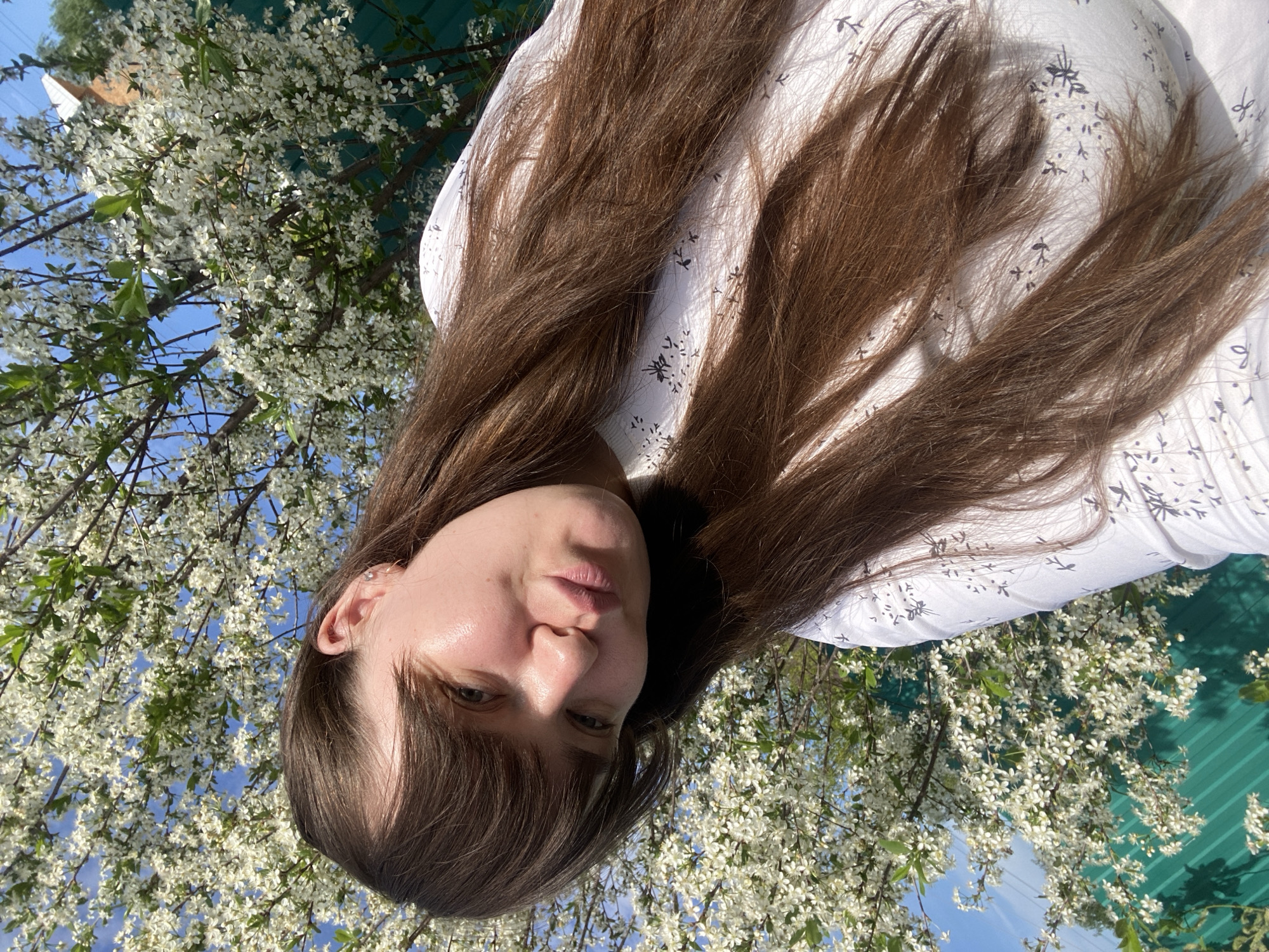A woman with long brown hair and bangs, wearing a white patterned shirt, is positioned against a backdrop of blooming white flowering trees under a blue sky. A green roof is partially visible. The location is suggested to be Votkinsk, Russia.