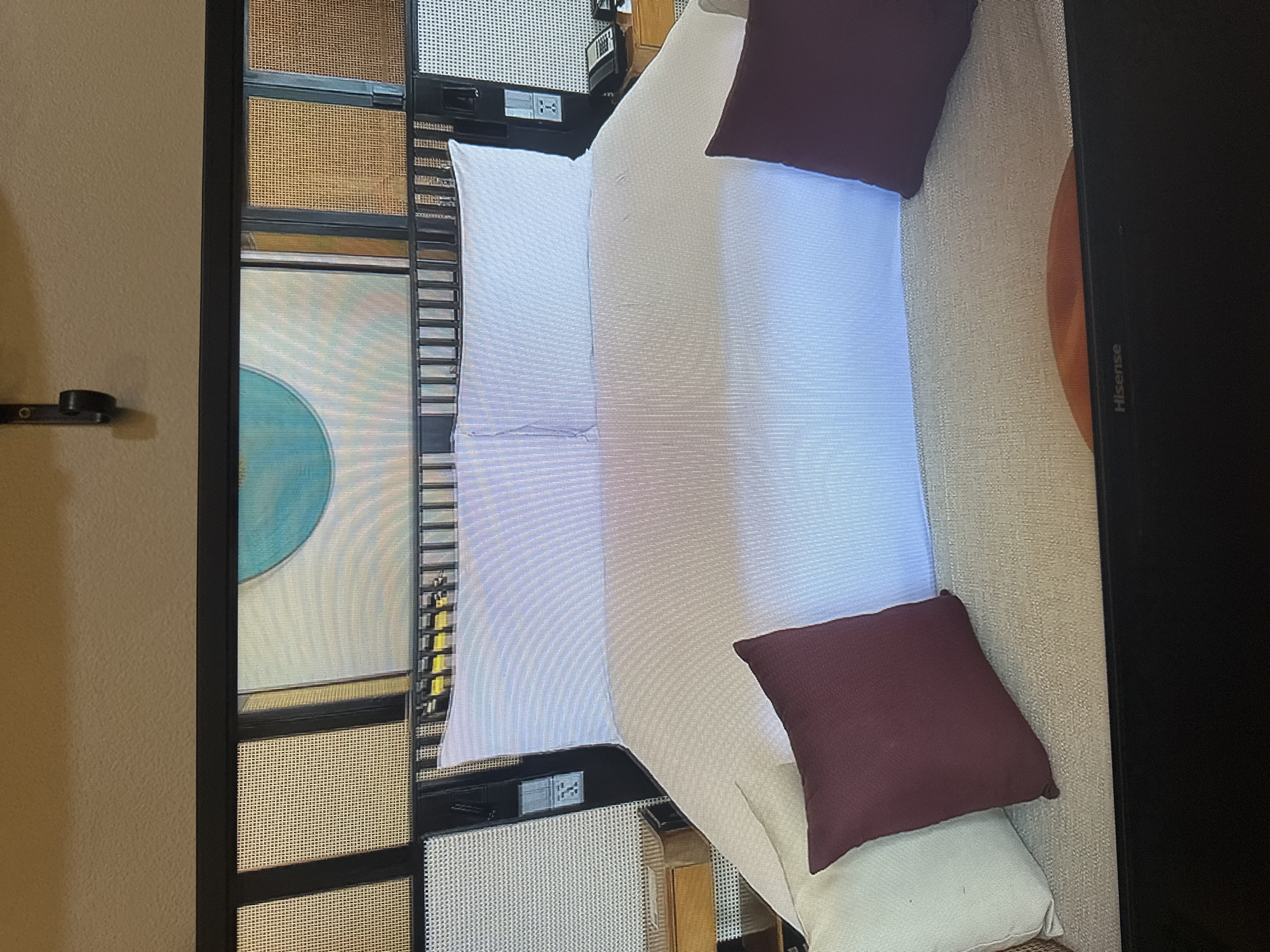 The image shows a close-up view of a bed in what appears to be a hotel room. The bed is covered with white bedding and has two large white pillows at the head. In front of the white pillows are two accent pillows, one light beige and one dark red. The headboard of the bed is made of dark wood with a lattice pattern and extends to the sides, where it frames a wall adorned with what looks like woven wall coverings and a large circular blue artwork. To the left and right of the bed, visible parts of nightstands can be seen. The nightstand on the left has a wooden drawer, and the one on the right has a black landline telephone and a wooden surface. Electrical outlets are visible on the wall next to the headboard. The bottom of the image shows a black Hisense television screen. There is no indication of the specific location in Kyiv, Ukraine, within the image itself.