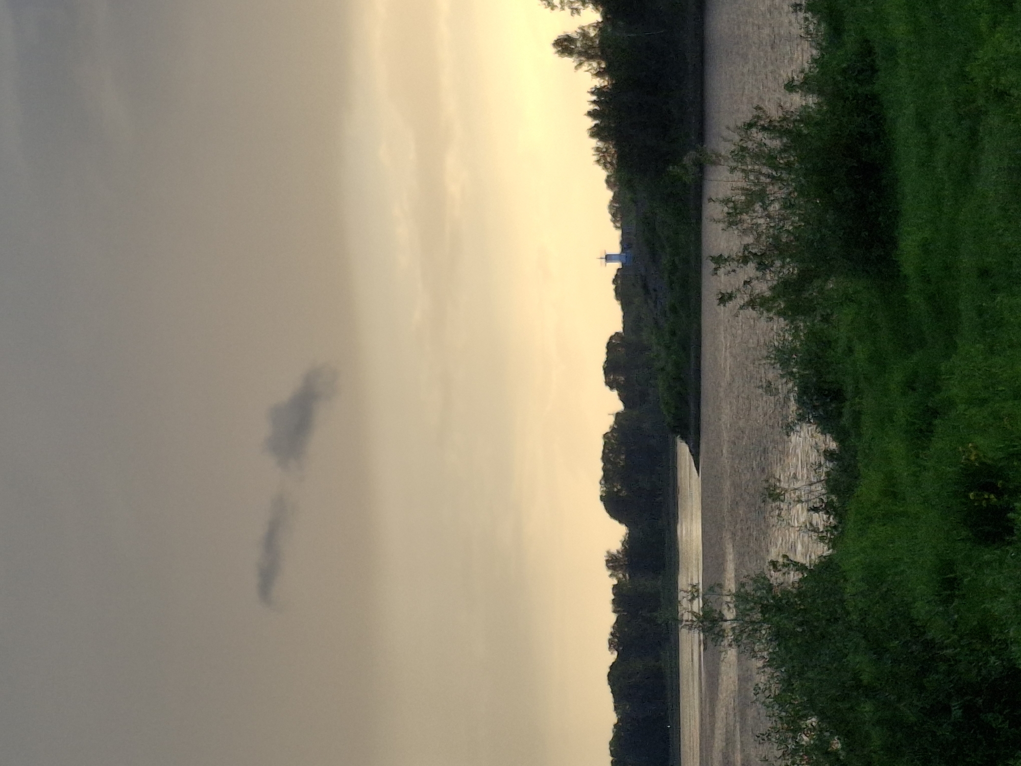 The image depicts a serene river scene at dusk, likely in Chaikovsky, Russia. In the foreground, lush green grass and dense foliage create a natural embankment leading towards the water. The river itself is calm, reflecting the soft, diffused light of the setting sun. Across the river, a line of dark trees forms a silhouette against the pale, hazy sky, which is characterized by muted tones of yellow and grey with a few faint clouds. Perched on a distant hill, a small white structure, possibly a lighthouse or a monument, stands out against the tree line, adding a focal point to the landscape. The overall impression is one of peacefulness and natural beauty.