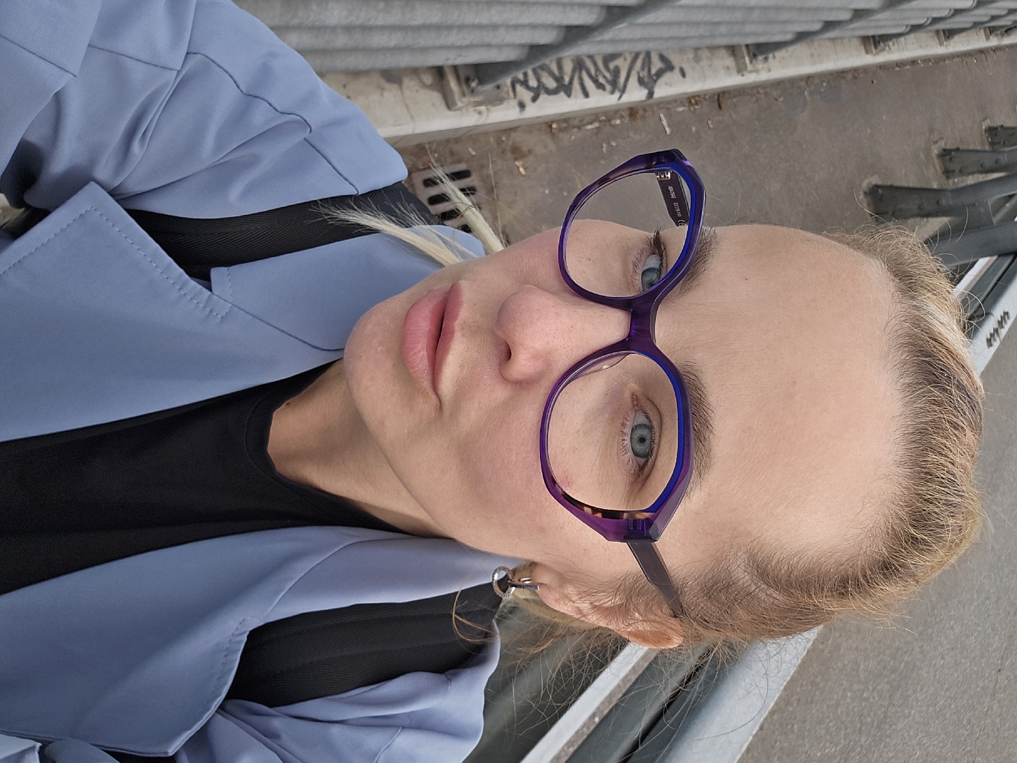 A Caucasian woman with blond hair pulled back and wearing blue-framed glasses is positioned in the foreground of the image. She is wearing a blue jacket over a black shirt. Her gaze is directed at the camera. In the background, there's a concrete surface with graffiti and metal railings. The setting appears to be urban, potentially a bridge or elevated walkway. No specific location within Sochi, Russia, can be determined from the image.