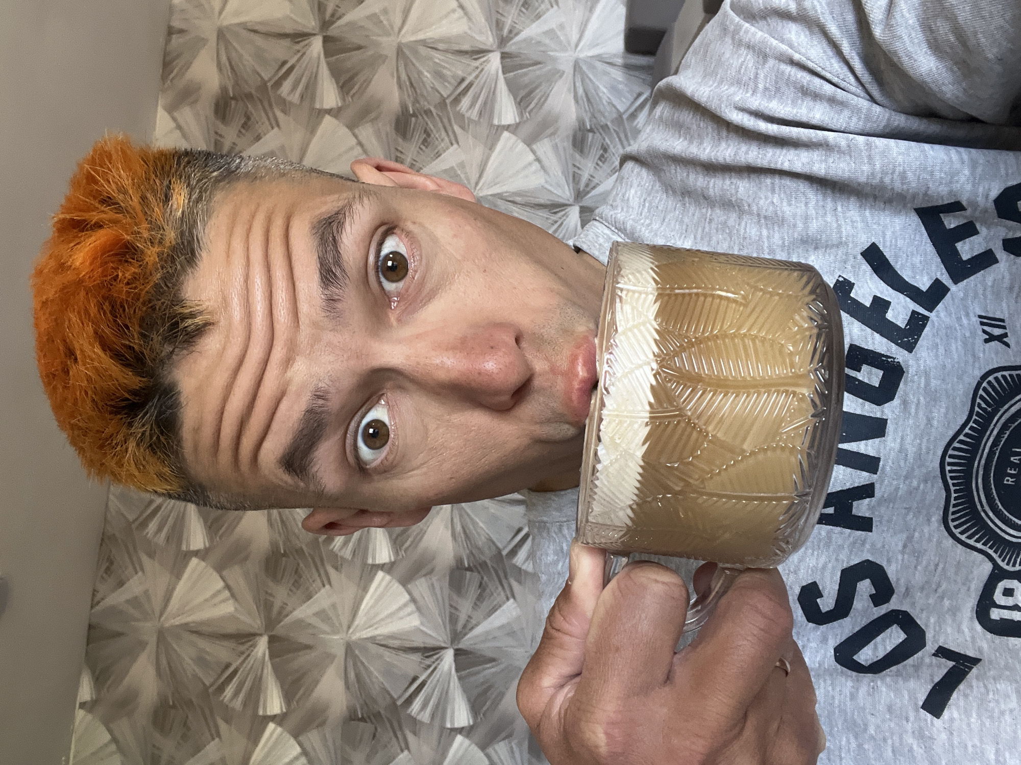 A middle-aged Caucasian man with orange-dyed hair, light skin, and a wrinkled forehead is holding a glass mug up to his mouth, possibly about to drink from it. His eyes are wide and he appears to be making a humorous expression. He is wearing a grey t-shirt with "LOS ANGELES" printed on it. The background consists of a patterned wallpaper in shades of grey and white. The specific location is not discernible from the image.