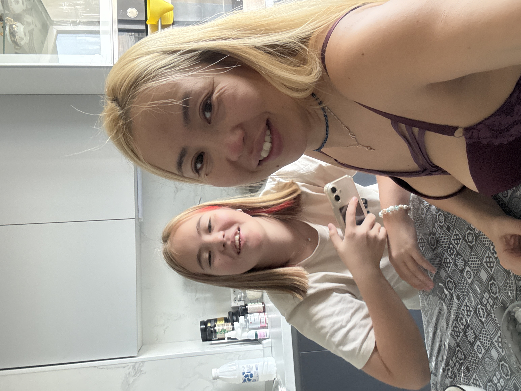 Two individuals are seated at a table in a kitchen. The person in the foreground, appearing to be an adult female with blonde hair, is looking at the camera and smiling. She is wearing a dark purple bra with decorative straps and a necklace. The second individual, appearing to be a younger female with blonde hair with red streaks, is seated behind her and to the left. She is also looking towards the camera, smiling, and holding a cell phone in her right hand. She is wearing a light-colored t-shirt and a bracelet. The kitchen appears modern with white cabinets and grey countertops. Various bottles and jars are visible on the countertop behind the younger individual. The table is covered with a patterned tablecloth.