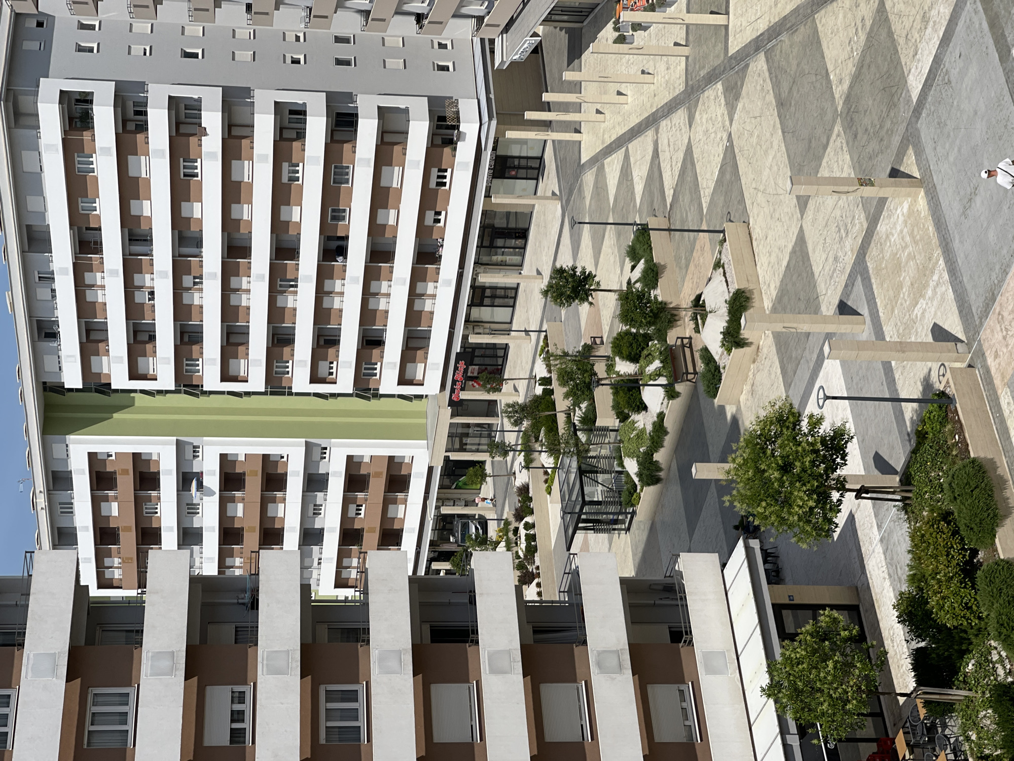 The image depicts a modern urban courtyard in Podgorica, Montenegro, enclosed by multi-story residential buildings. The courtyard features a checkered paving pattern with large square stones. Vertical, rectangular pillars are strategically placed throughout the space. Greenery is incorporated through planters with bushes and small trees, as well as larger trees with visible trunks. A bench is visible in one of the planted areas.
The buildings surrounding the courtyard have balconies and white shutters on their windows. One building prominently displays a vertical green facade. At street level, commercial spaces with storefronts are integrated into the buildings, including a shop with signage that appears to read "pierre cardin."
In the foreground, a person wearing a white cap and a white shirt is walking away from the camera. The lighting suggests it is daytime, with shadows cast by the buildings and pillars, indicating a sunny environment. The overall impression is one of a newly developed, clean, and contemporary urban living and commercial space.