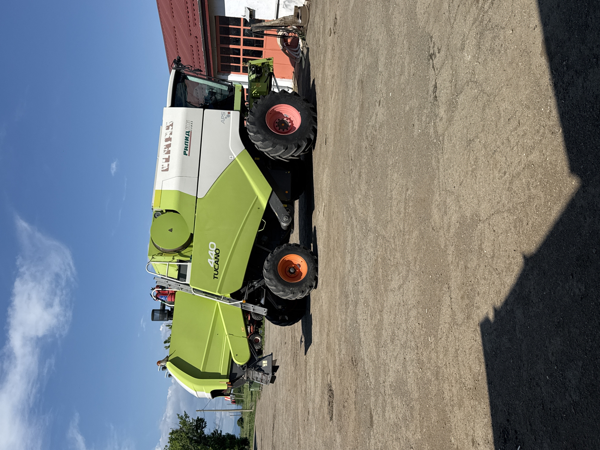 The image shows a Claas Tucano 440 combine harvester parked on a concrete surface. The harvester is green and white, with large tires and a header attached. The sky is blue with some clouds visible. A building with a red roof is partially visible behind the harvester. The image was taken in Sarnevo, Bulgaria.