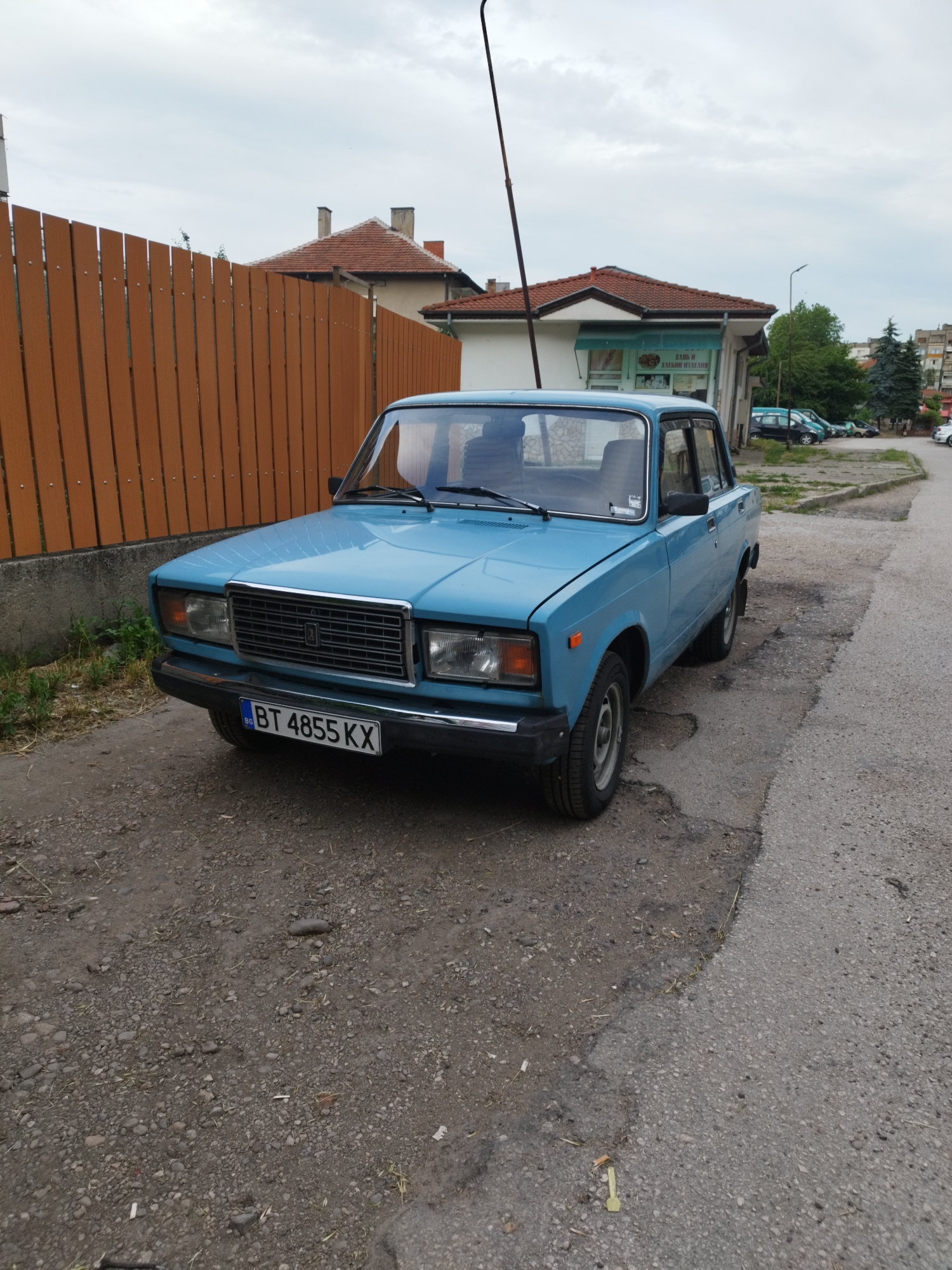 The image shows a blue Lada car parked on a street in an urban area. The car has a Bulgarian license plate that reads "BT 4855 KX". The street is partially paved and partially covered with gravel. Behind the car is a wooden fence and a building with a red tile roof. Other buildings and trees are visible in the background. The sky is overcast. The location is in the city of Unknown, Unknown.
