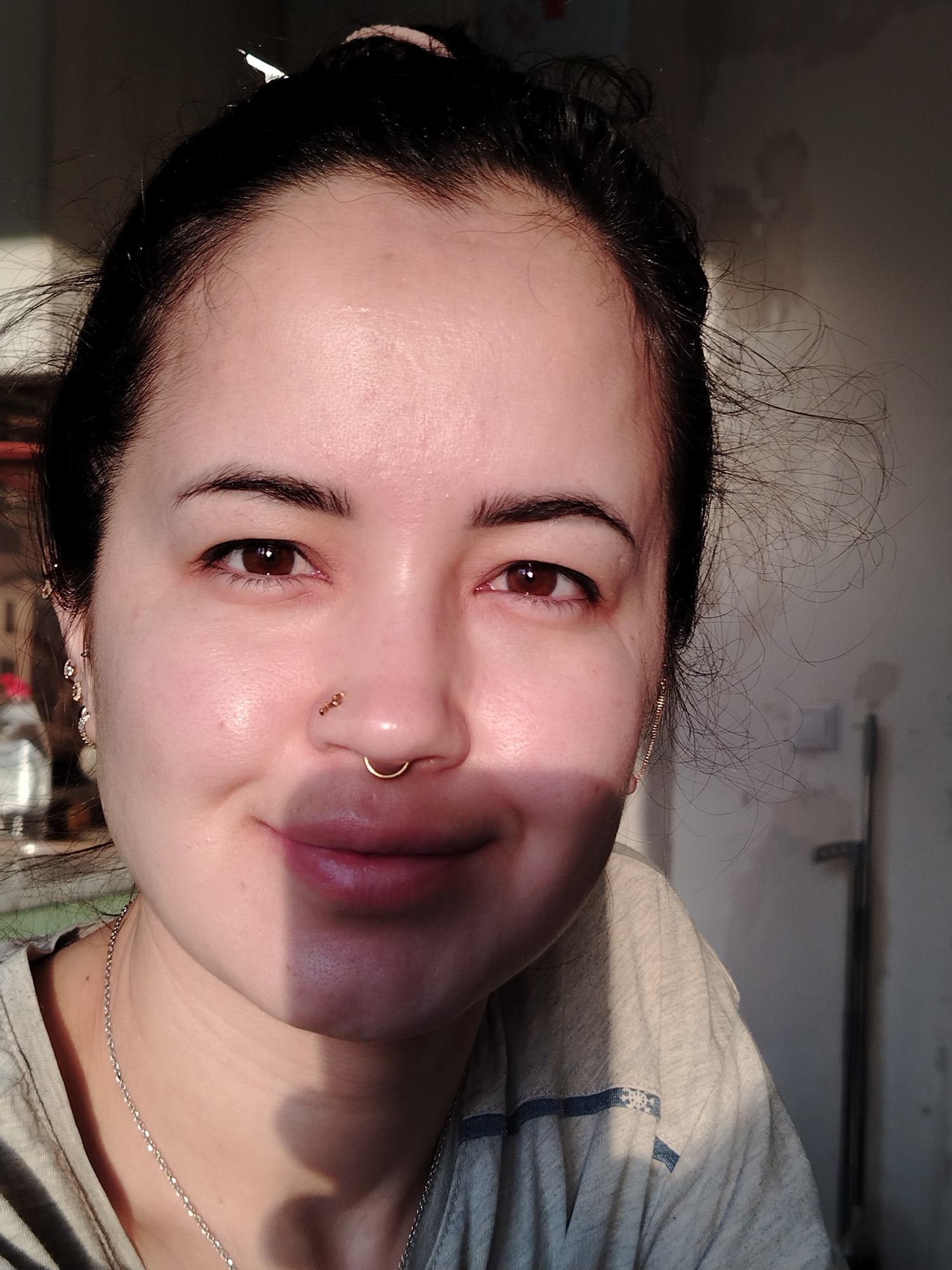 A close-up, indoor portrait of a light-skinned woman with dark hair pulled back. She has brown eyes, a nose ring, and is wearing a silver necklace and earrings. The woman is smiling slightly. The right side of her face is in shadow. She is wearing a light-colored t-shirt. The background is out of focus, showing a partially visible wall, door and blurry shapes which are indiscernible. There is no visible information to suggest the location is Noviy, Russia.