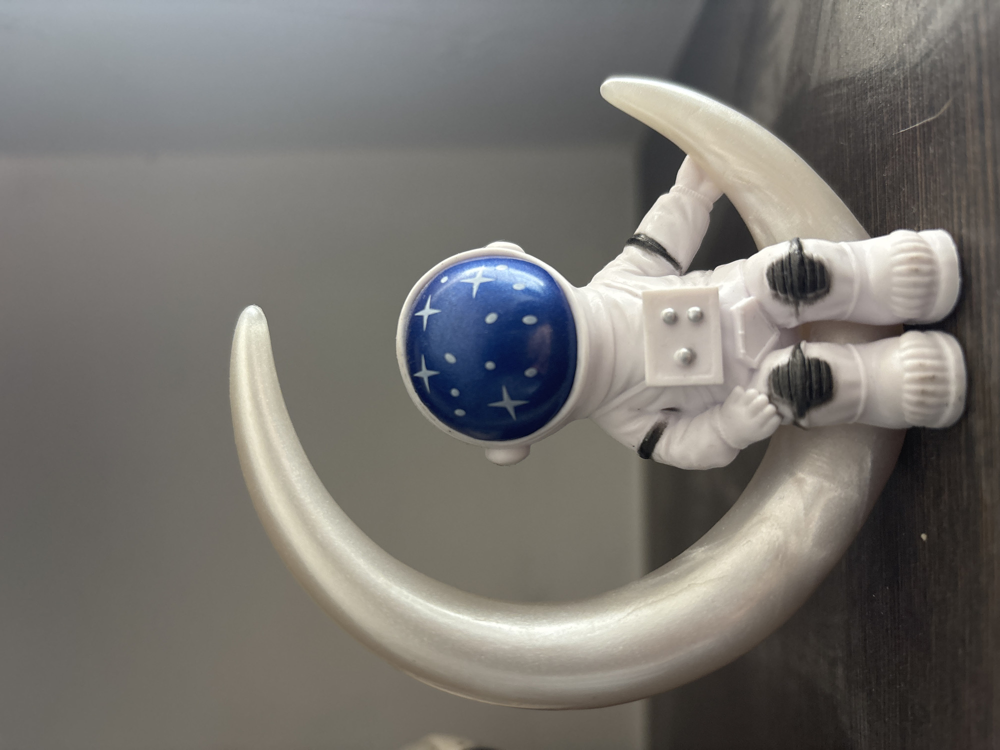 A close-up studio shot features a white, plastic astronaut figurine reclining on a crescent moon. The astronaut has a blue helmet adorned with white stars and dots, and black detailing on the gloves and boots. The crescent moon is light gray. The background is gray on the left and brown on the right. The location is Jesús del Monte, Mexico, but this is unverifiable from the image.