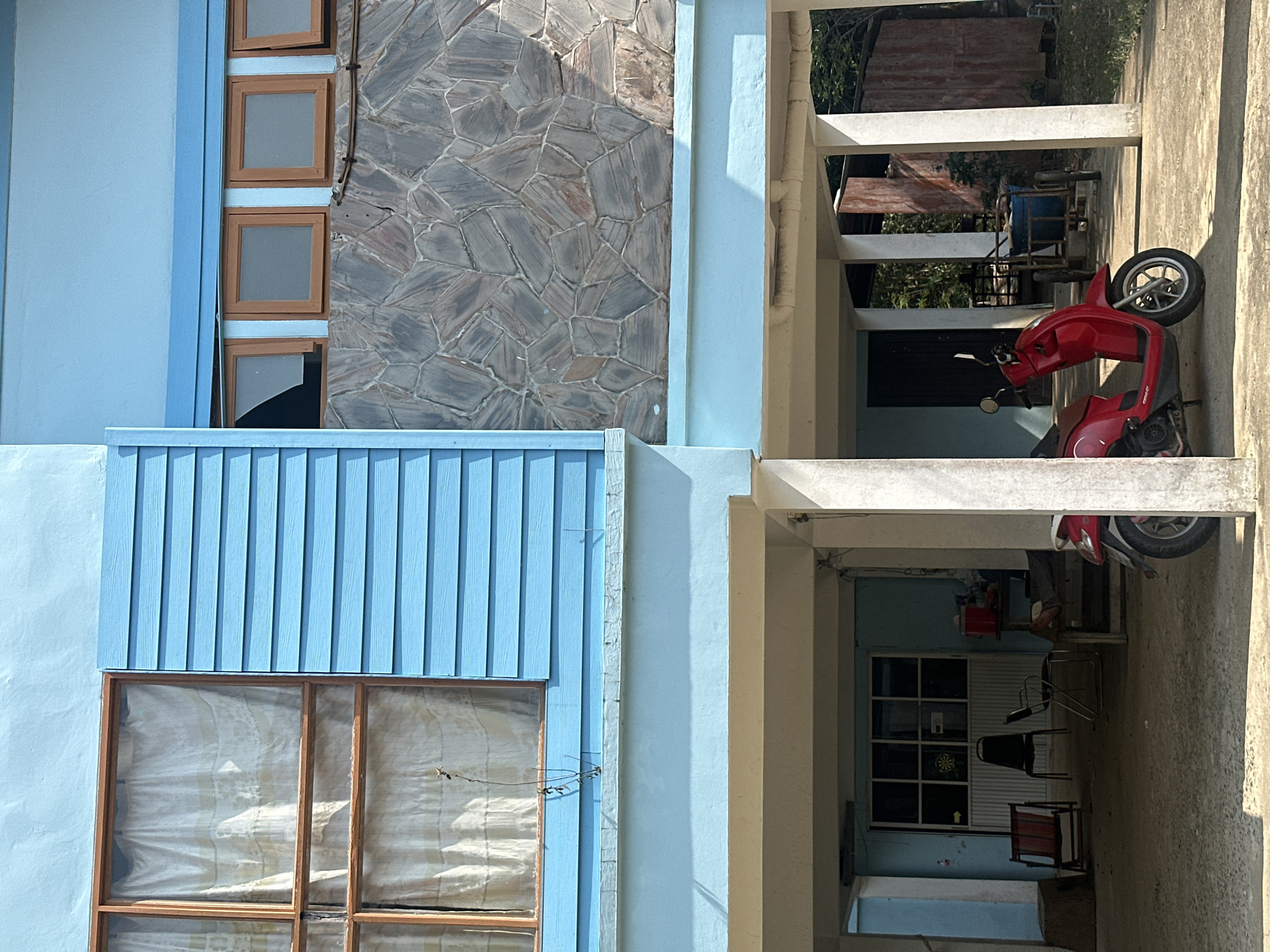 The scene is an exterior shot of a building in Ao Hin Khok, Thailand. The building has light blue siding and a stone wall facade. There are several windows, some with curtains visible inside. A red scooter is parked under a covered walkway supported by cream-colored pillars. Inside the walkway, there are chairs, a table, and what appears to be a dartboard hanging on the wall.