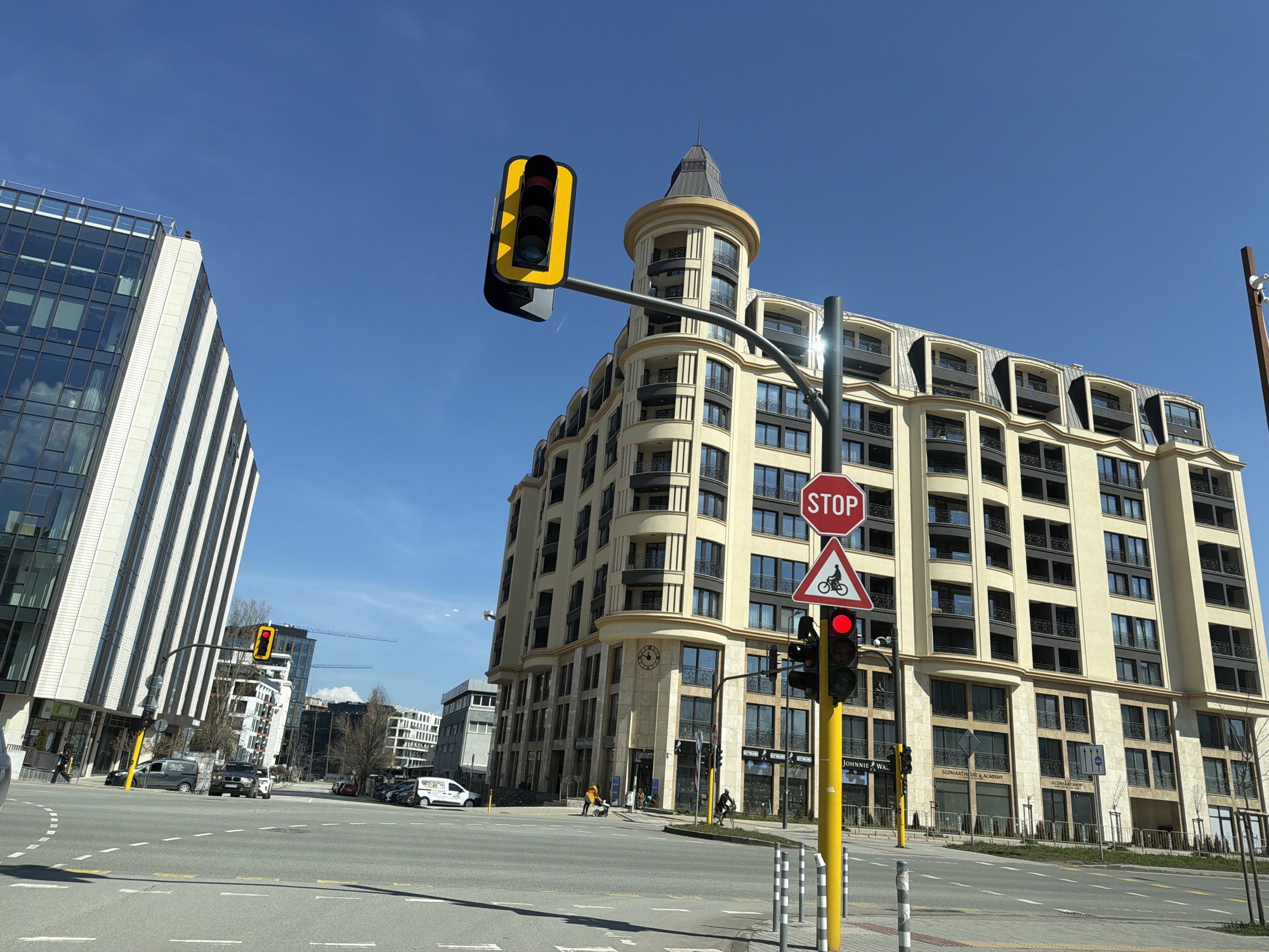 The image depicts a street scene in Sofia, Bulgaria, characterized by a mix of modern and classically styled buildings under a clear blue sky. A tall, light-colored building with a tower-like structure dominates the right side of the frame. Its architecture features multiple floors with numerous windows and balconies. To the left, a modern glass-clad building with alternating white stripes is visible.
In the foreground, a traffic light pole displays a stop sign and a bicycle crossing warning sign above a red traffic light. Various vehicles are present on the road, including cars and vans. Pedestrians are observed on the sidewalks near the buildings. The road is marked with white lines, and several utility poles are placed along the sides of the street.