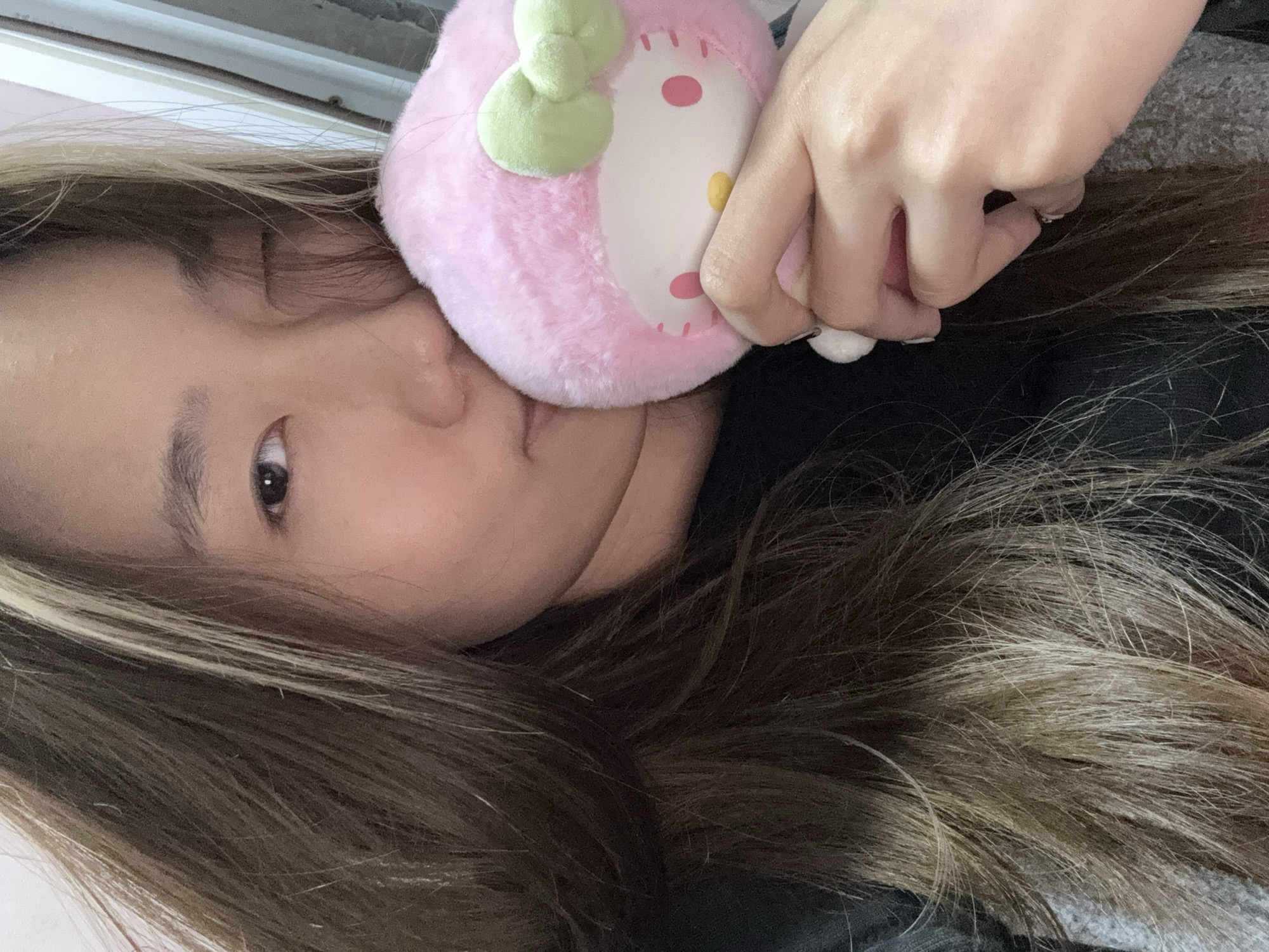 A woman with brown and blonde highlighted hair lies on her side, holding a pink Hello Kitty plush toy near her face. She is wearing a dark-colored shirt. The background is blurred, but shows a light-colored wall or surface. There is no information to confirm that this image was taken in Shapingba District, China.