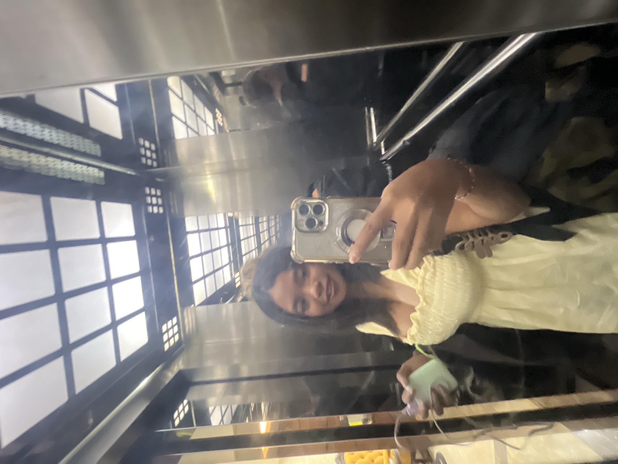 A young woman is taking a selfie in a mirrored elevator. She is wearing a pale yellow, smocked top and has dark hair. She is holding a light green object in her left hand and her phone in her right hand. Reflections of the elevator ceiling, which has a grid of lights, and other people are visible in the mirrors. The elevator appears to be in a modern building.
