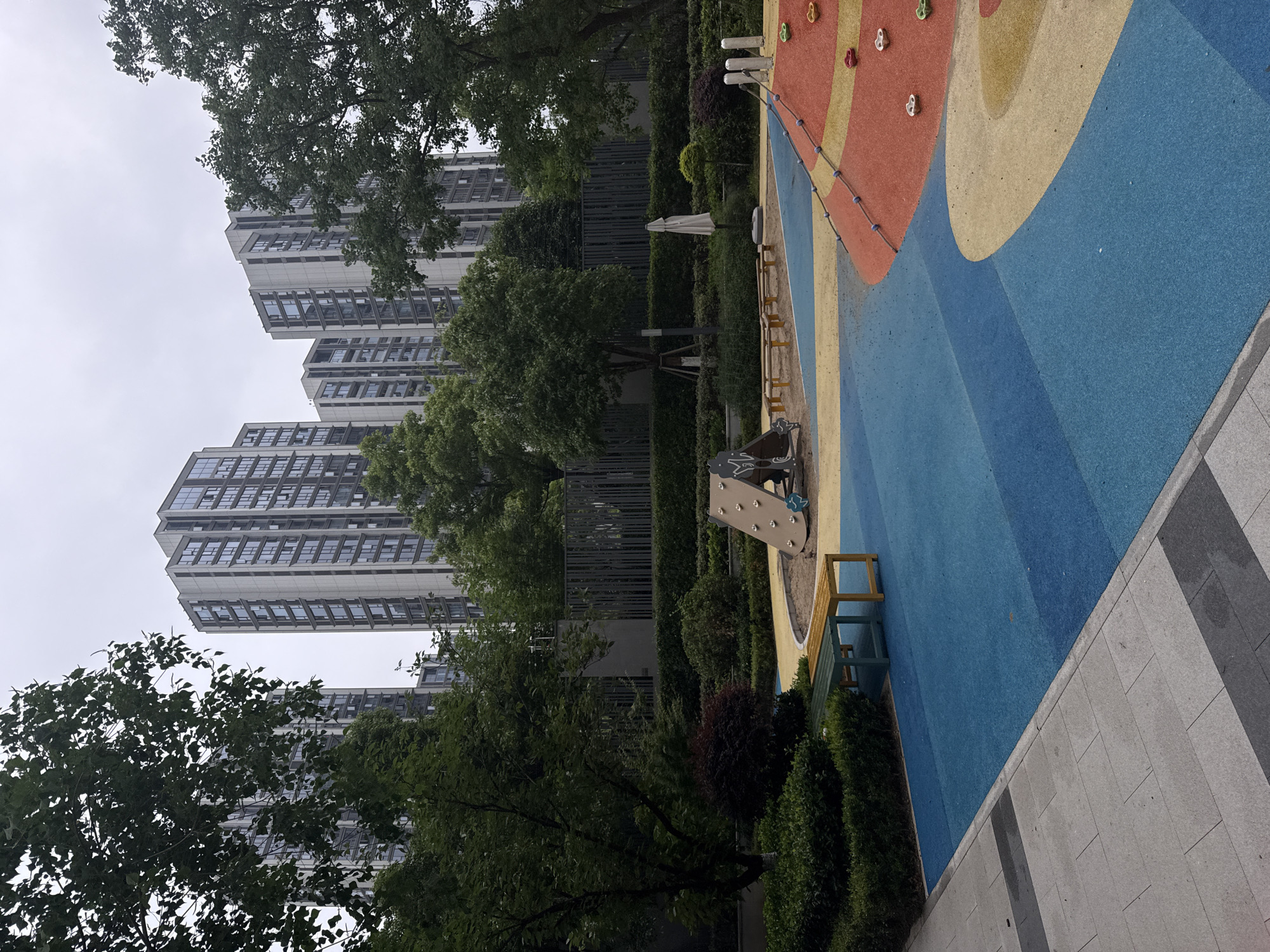 This image captures an outdoor scene in Yiwu, China, featuring a modern children's play area set against a backdrop of tall residential buildings and abundant greenery.
The foreground is dominated by a vibrant, multi-colored playground with a soft, padded surface. Sections of bright blue, yellow, and red are prominent, with abstract shapes creating a dynamic visual. A small climbing structure, possibly a wall, with various handholds (red, green, white/gray) is integrated into the play surface. Adjacent to the play area, a sandpit is visible, along with several benches—one made of wood and another appearing to be green-painted metal. Paved walkways, constructed from light gray and darker gray rectangular tiles, border the playground on the right side.
Behind the play area, a thick line of neatly trimmed green hedges and several mature trees with dark green foliage create a natural buffer. Through and above the trees, multiple high-rise apartment buildings with contemporary architecture are visible. These buildings feature numerous windows and light-colored facades, towering under an overcast sky. A dark fence or barrier runs behind the hedges, separating the recreational space from the buildings. A white, folded umbrella and a small round bush are also seen behind the hedges.
The scene appears tranquil and devoid of people, suggesting a quiet moment in the recreational space. The weather is cloudy, with a bright, diffused light indicating an overcast day. There is no visible text in the image.
