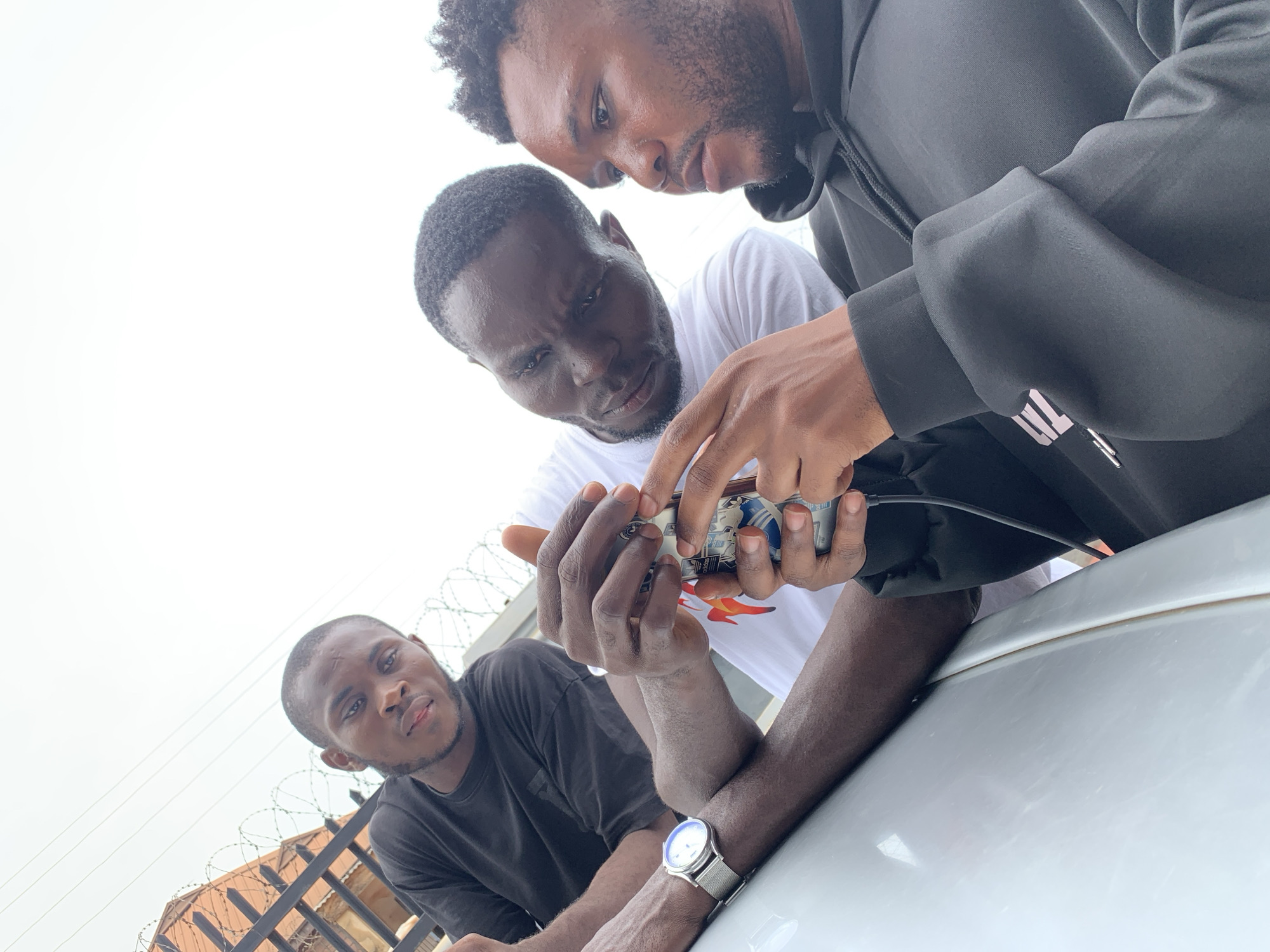 Three young men are depicted in an upward-angled outdoor shot, set in Ikorodu, Nigeria. Two of the men, positioned centrally and to the upper right, are intently focused on a small, cylindrical electronic device they are holding. The man in the black hoodie, on the right, points at a component on the device, while the man in the white t-shirt looks closely at it, suggesting they are inspecting or collaboratively working on the item. Wires and connectors are visible on the device.
The third man, in a black t-shirt, leans in from the lower left, looking towards the camera with a neutral expression. He wears a silver-colored watch with a mesh band on his left wrist. The men appear to be leaning on a white car or vehicle surface, which occupies the lower right portion of the frame.
In the background, under a bright, overcast sky, a metal fence topped with barbed wire is visible, along with parts of a building featuring a reddish-brown roof, indicating an urban or suburban setting. The man in the black hoodie wears a white logo, possibly Adidas's three parallel bars, on his sleeve. The white t-shirt features a graphic with red and orange flame-like designs, though specific text is unreadable. The scene takes place during the daytime, with an atmosphere of focused attention and collaborative activity.