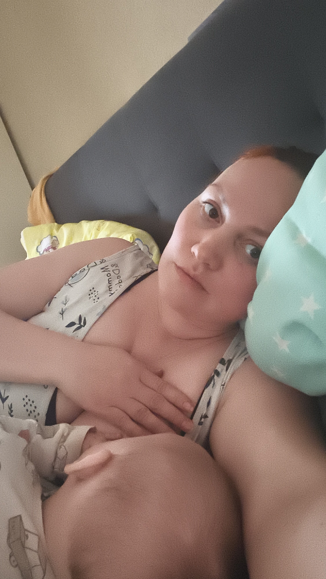 A woman is lying down, apparently breastfeeding a baby. She has fair skin, reddish hair, and is looking directly at the camera. She is wearing a patterned tank top. Her hand is on her chest. The baby's head is visible, nestled against her. They are covered with a patterned blanket. A blue pillow with white stars is behind her head, and a gray headboard is behind the pillow. There is a yellow object in the background as well. The location is Unknown, Unknown.