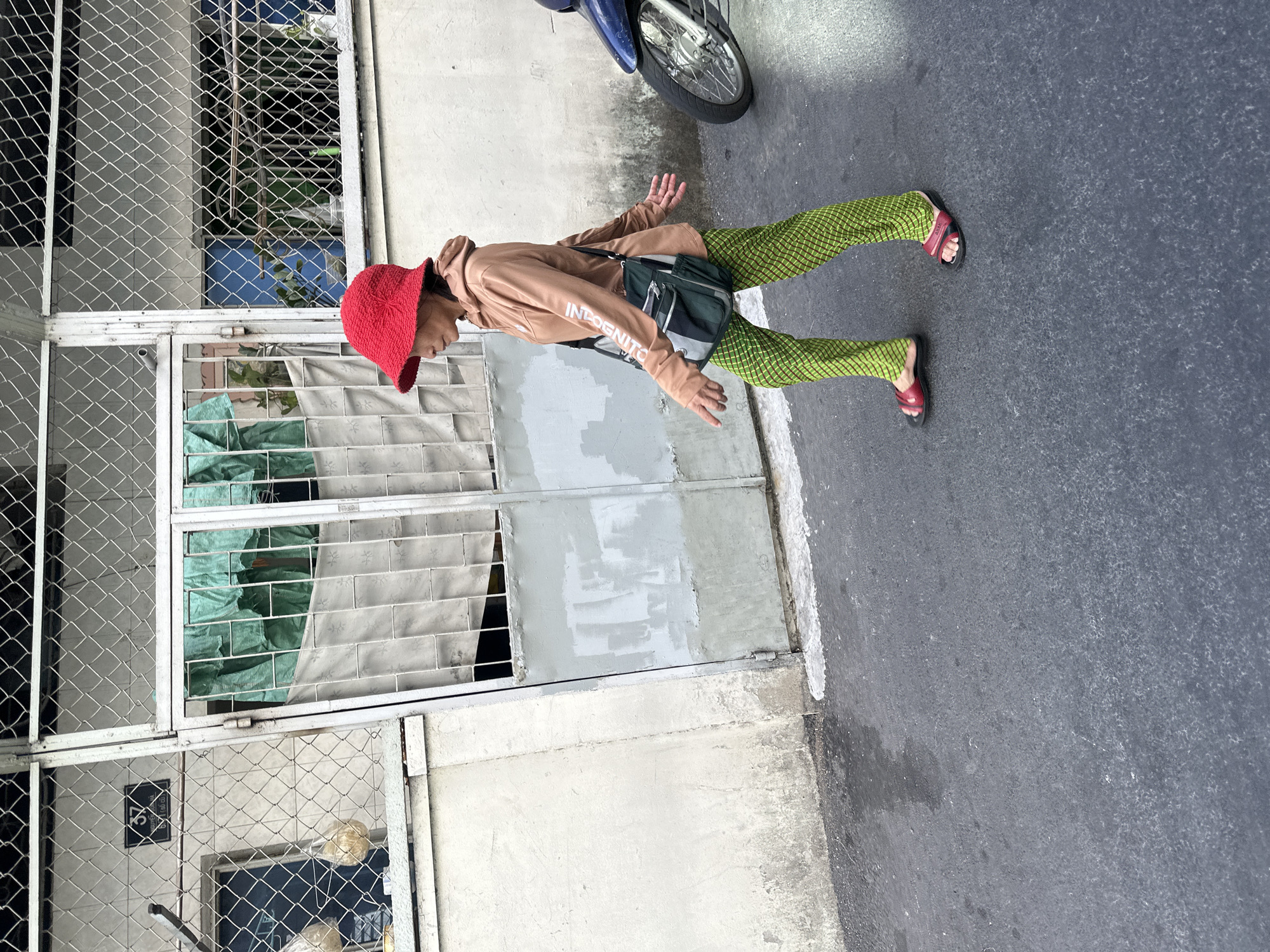 An individual is standing on a street, likely in Thủ Đức, Vietnam. The person is wearing a red bucket hat, a light brown hooded sweatshirt with the word "INDOGNITO" printed on the sleeve, green patterned pants, and red sandals. They are carrying a green and gray shoulder bag. The individual stands to the left of a closed metal fence with a wire mesh structure. The street is gray asphalt. A parked motorcycle is visible in the upper right corner of the image. The weather appears to be overcast.
