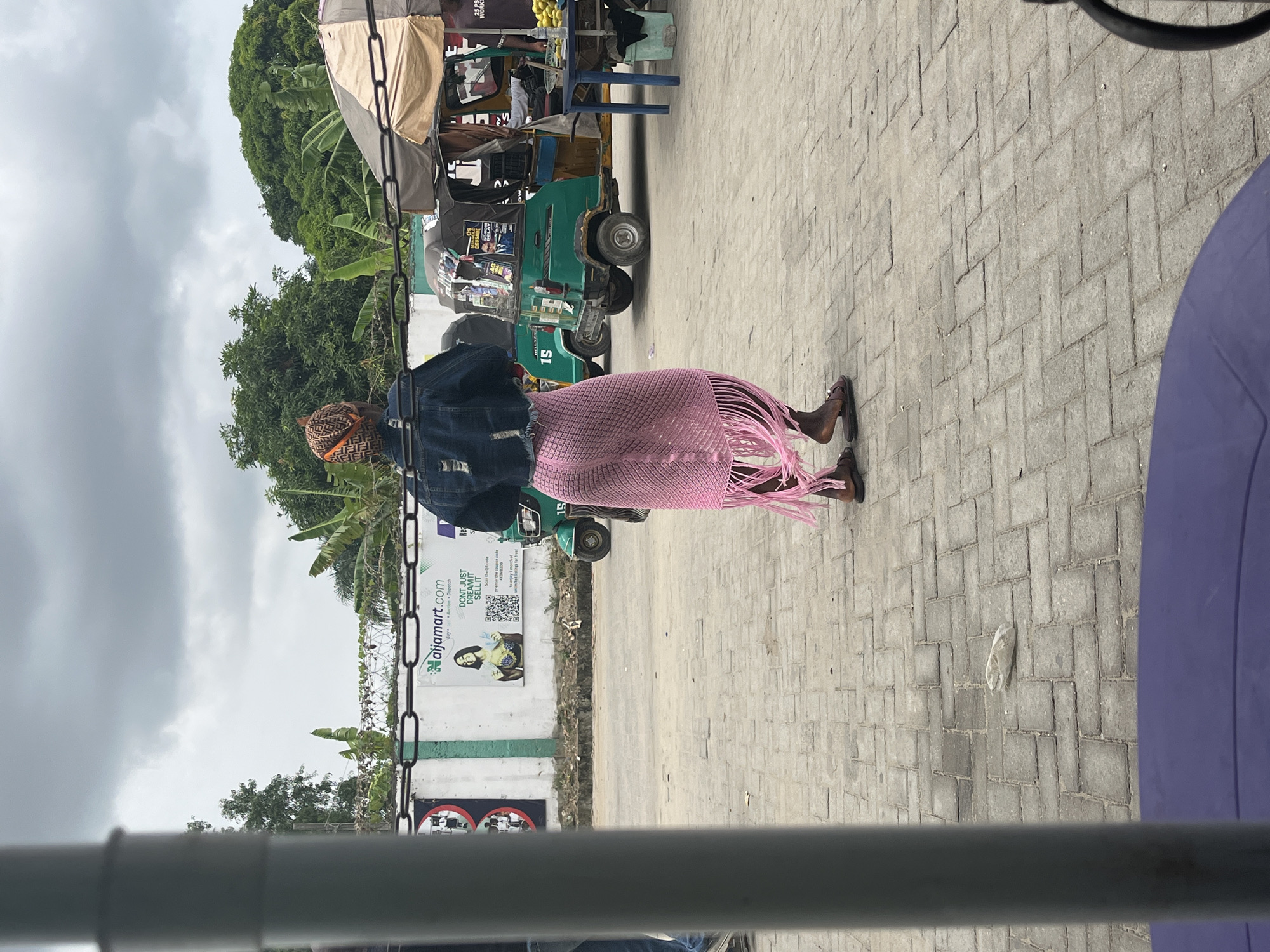 The scene is set in Lekki, Nigeria. The image focuses on an elderly woman, her back to the camera, wearing a denim jacket and a pink fringed skirt. She is walking along a paved pathway. To her left, there is a wall with an advertisement for "aijamart.com" and some other signage. In the background, there's a green auto-rickshaw and a fruit stand. The sky is overcast with heavy clouds. A piece of trash lies on the pathway. The image seems to have been taken from a moving vehicle, with parts of the vehicle's frame visible in the foreground.
