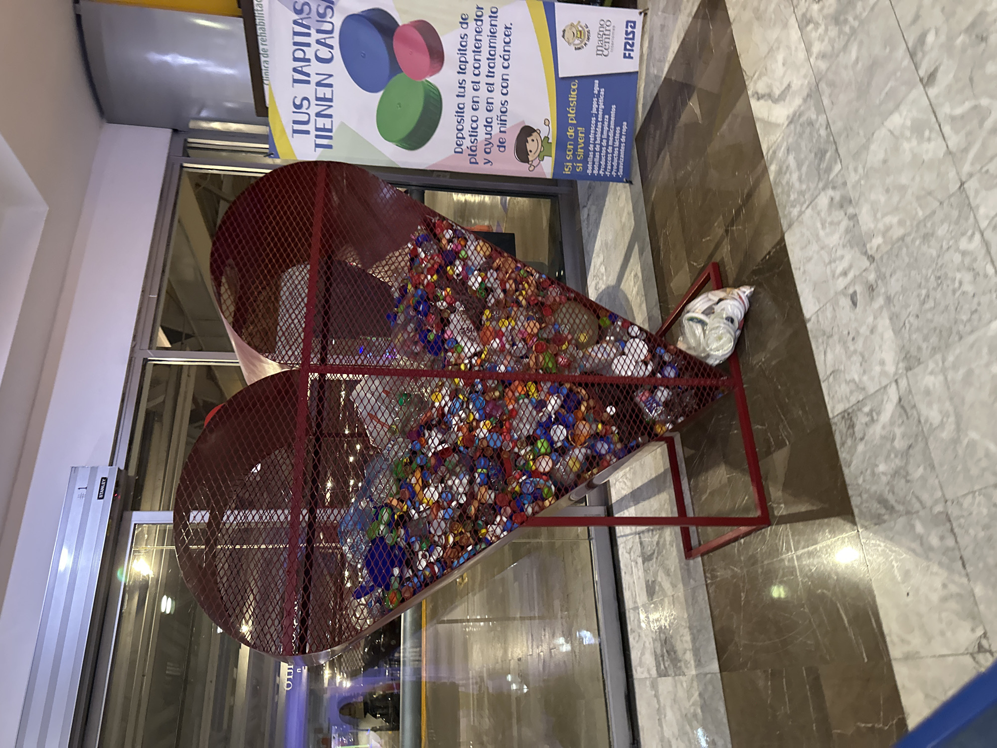 The image captures an indoor scene featuring a large, heart-shaped container filled with colorful plastic bottle caps. The container is made of red metal wire mesh and sits on a matching red metal frame. Adjacent to the container, a promotional banner is displayed, which states "TUS TAPITAS TIENEN CAUSA" (Your caps have a cause), encouraging the donation of plastic bottle caps for the treatment of children with cancer. The banner lists acceptable plastic items, including bottles of soda, juice, water, energy drinks, cleaning products, medicine containers, dairy products, and fabric softeners. A bag filled with white plastic caps is positioned beside the heart-shaped container. The container is located inside the magno Centro Frisa in the city of Huixquilucan de Degollado, Mexico. The floor is tiled with a marble-like pattern in shades of gray and brown. Through the glass doors, blurred lights and reflections suggest an exterior or another interior space. A "STANLEY #1" sign is affixed to a window frame.