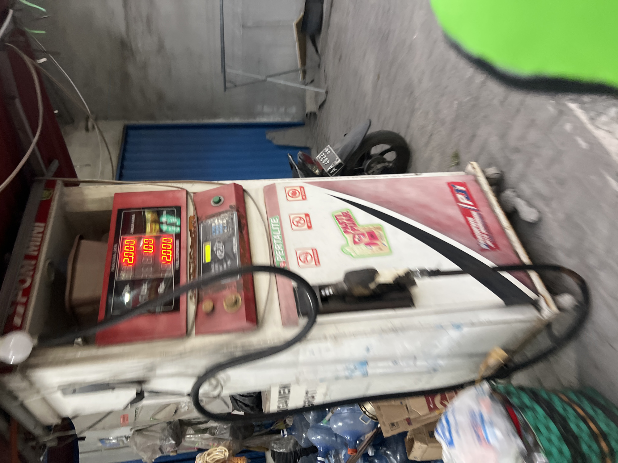 A red and white gasoline pump is the central focus of the image, located in Sendangadi, Indonesia. The pump displays red digital numbers that read "12.000" on three separate lines. A black hose is coiled around the pump. To the left of the pump, a stack of large blue water jugs is visible, along with some cardboard boxes and a green woven basket filled with plastic bags. In the background, a blue folding door and a concrete wall are present. A motorcycle with a license plate that reads "AA 4022" is parked to the right of the gasoline pump. The ground appears to be a concrete or asphalt surface. The overall scene suggests a commercial or retail setting, possibly a small convenience store or gas station.