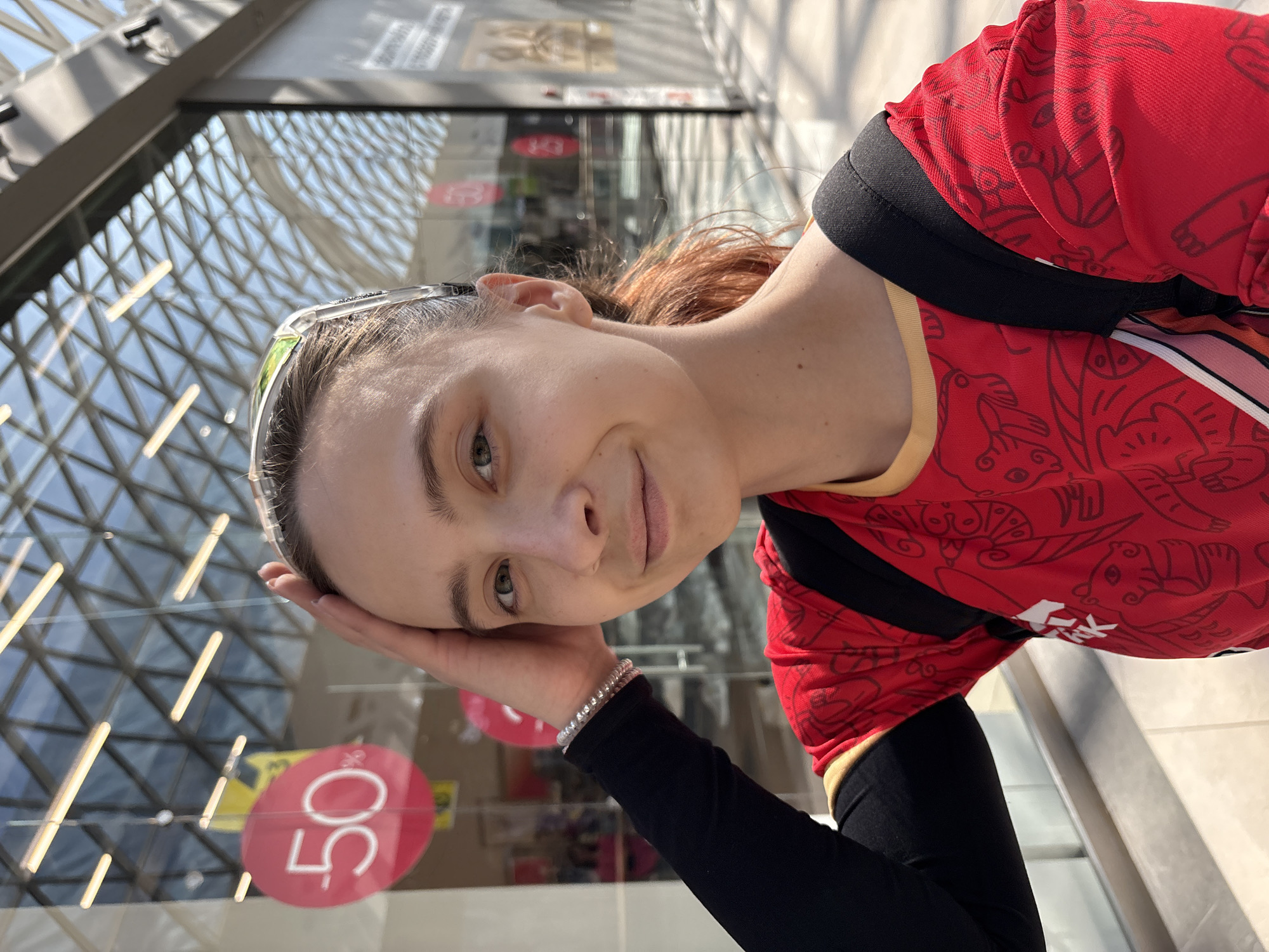 A woman with reddish-brown hair is seen from a low-angle selfie perspective inside what appears to be a shopping mall. She is wearing a red shirt with dark outlines and black long sleeves with a bracelet on her left wrist. She rests her head on her left hand. Behind her is a glass wall with a 50% off sign and the architectural dome-like structure of the shopping mall is visible. The woman appears to be in the city of Perm, Russia, based on the design of the dome.