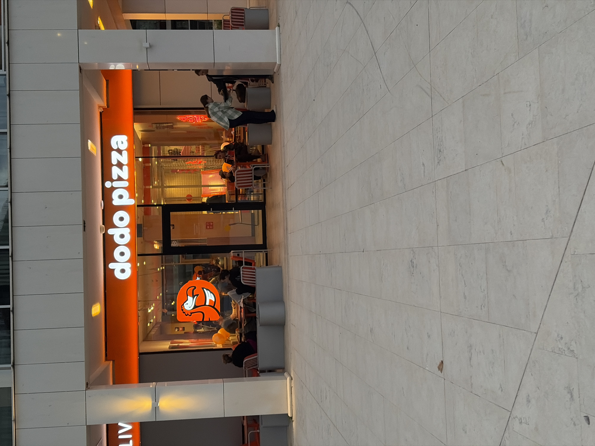 The image depicts the exterior of a Dodo Pizza restaurant. The restaurant's storefront is predominantly orange with the "dodo pizza" logo displayed vertically. People are visible inside the restaurant. Some are seated at tables. Others are standing near the entrance. One man in a plaid shirt is outside the restaurant. The exterior of the building is primarily white or light gray. The ground is paved with large, light-colored tiles. Based on the Dodo Pizza branding and online presence, the location could be in Budva, Montenegro.