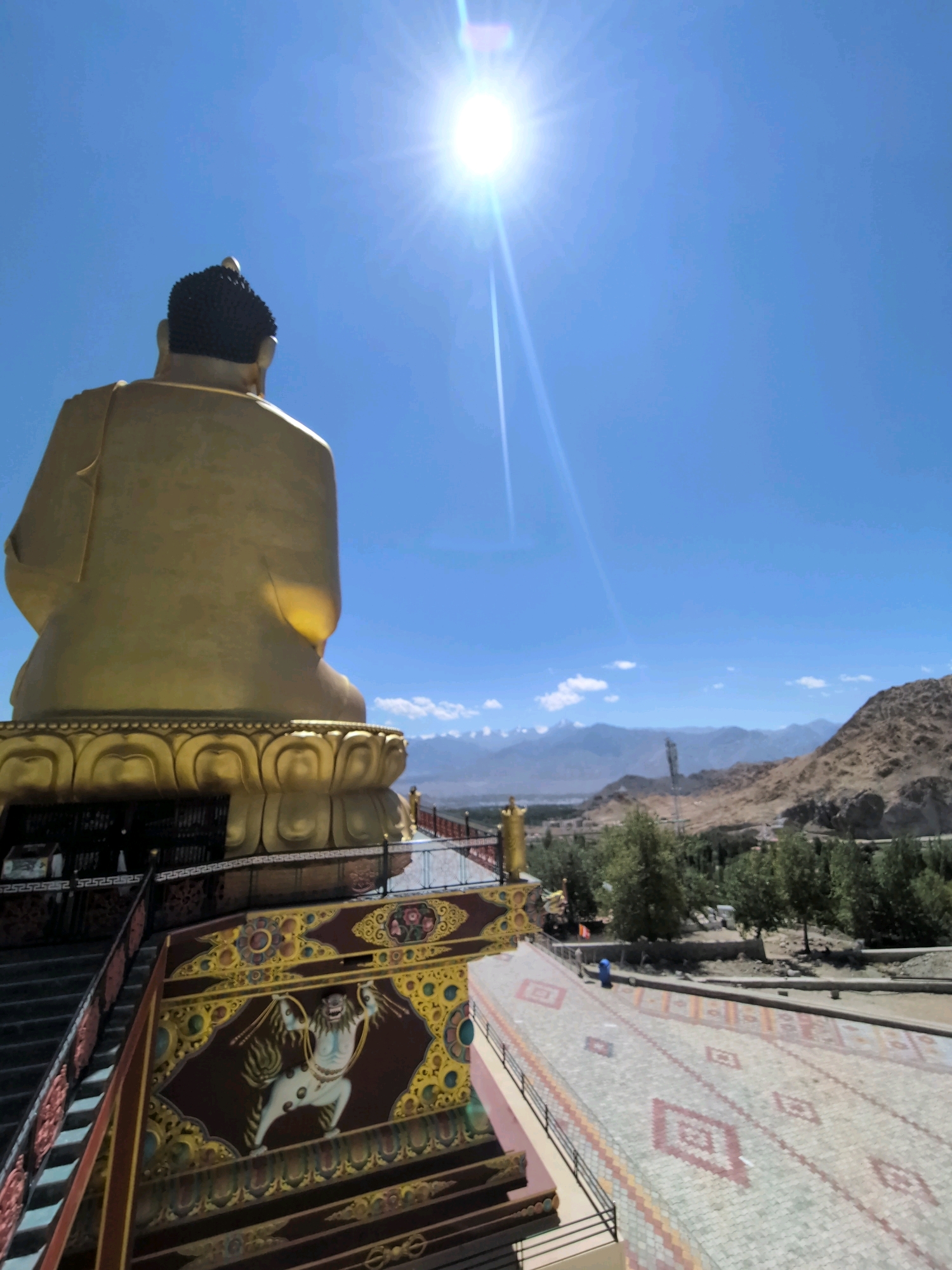 The image shows a view from behind a large, golden statue of Buddha, looking out over a landscape. The statue is seated on a decorative base, which is part of a larger structure with ornate carvings and paintings visible on its side. A stairway with red railings leads down from the statue's base. In the foreground, a paved walkway with a patterned design is visible. The background features a valley with trees and buildings, leading to a range of mountains under a bright blue sky with a prominent sun and lens flare. The scene is likely located in Leh, India, given the distinctive architecture and mountainous terrain often associated with Buddhist monasteries in the region.