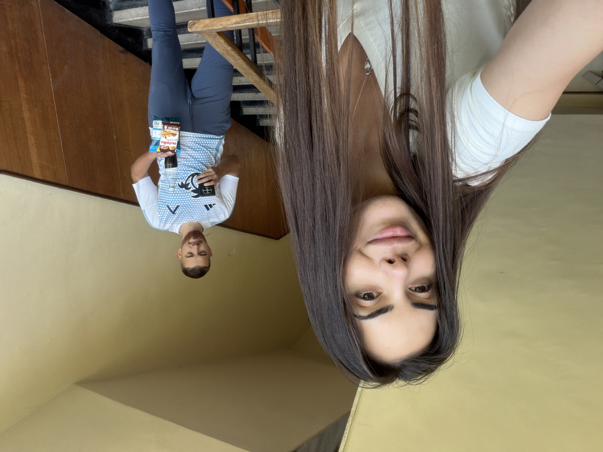 The image depicts two individuals in what appears to be an indoor setting, possibly a building staircase in Sofia, Bulgaria, given the likely origin. One person, possibly taking a selfie, occupies a significant portion of the frame, focusing on their face and long hair. They are wearing a light-colored top. The second person is positioned further back and appears to be holding items including a water bottle and a Nutella snack. They are wearing a blue and white athletic shirt and dark pants. The background shows stairs and wood paneling, indicating an interior space. The overall lighting is somewhat diffused.