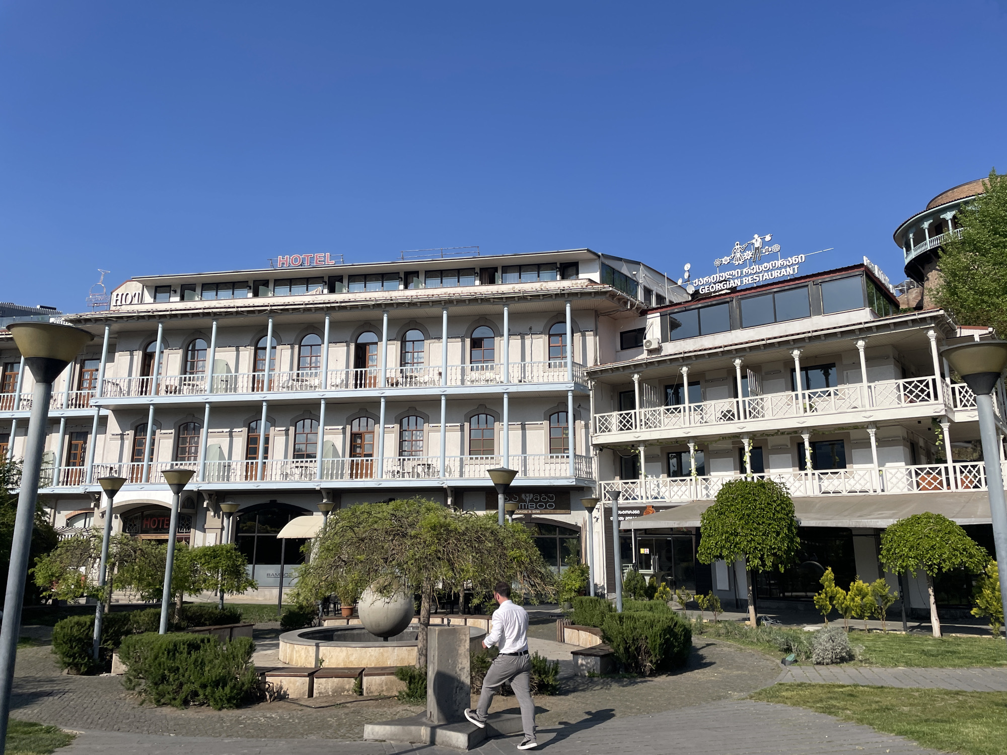 The image shows a street scene in Tbilisi, Georgia, featuring two adjacent buildings: a hotel and a Georgian restaurant. The hotel has multiple floors with balconies and a "HOTEL" sign on the roof. The Georgian restaurant, also with balconies, is topped with a sign in Georgian and English. A man in a light-colored shirt and grey trousers is walking through a landscaped area with a fountain. Street lamps and greenery are visible in the foreground. The sky is clear and blue.