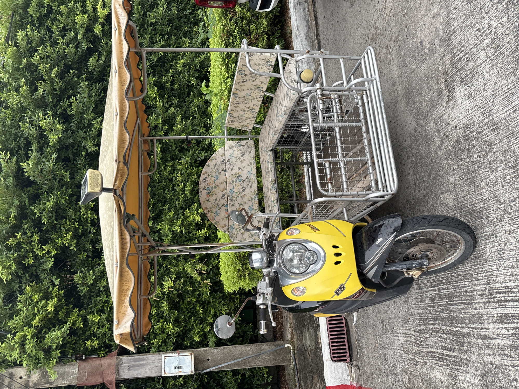 A yellow and black scooter is attached to a sidecar with a yellow canopy in Pattaya City, Thailand. The sidecar is made of metal and has a patterned fabric seat. Behind the scooter and sidecar, there is a dense green hedge. A concrete utility pole with electrical components is to the left of the scooter. The ground is paved with concrete.
