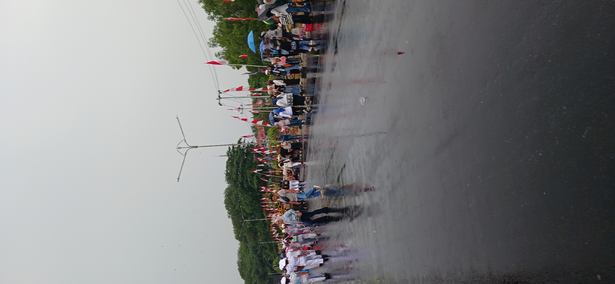 The image captures a street scene in Tanjung Balai Karimun, Indonesia, on a cloudy day, possibly after rain, as the asphalt surface is wet and reflective. A procession or parade is underway, marked by a line of people on the right side of the road and a group of individuals walking on the wet street on the left. Many of the participants, especially on the left, are wearing white uniforms, suggesting a school or organizational group. Indonesian flags, red and white, are prominently displayed along the procession route, mounted on poles and attached to vehicles.
In the foreground, the wet asphalt reflects the overcast sky and the figures walking on it. Two men are in motion on the road: one in a blue shirt and jeans, and another in a light-colored shirt and dark pants. The man in blue appears to be walking forward with his right leg extended.
Further down the road, a larger group of people are gathered, some standing and observing the procession. Several individuals are holding umbrellas, one blue and one black, indicating the possibility of ongoing or recent rain. The crowd is a mix of adults and possibly children.
Behind the procession, a line of trees forms a backdrop, and beyond that, buildings are partially visible. A street lamp with a distinctive design stands tall in the middle ground. The overall atmosphere suggests a community event or celebration.
