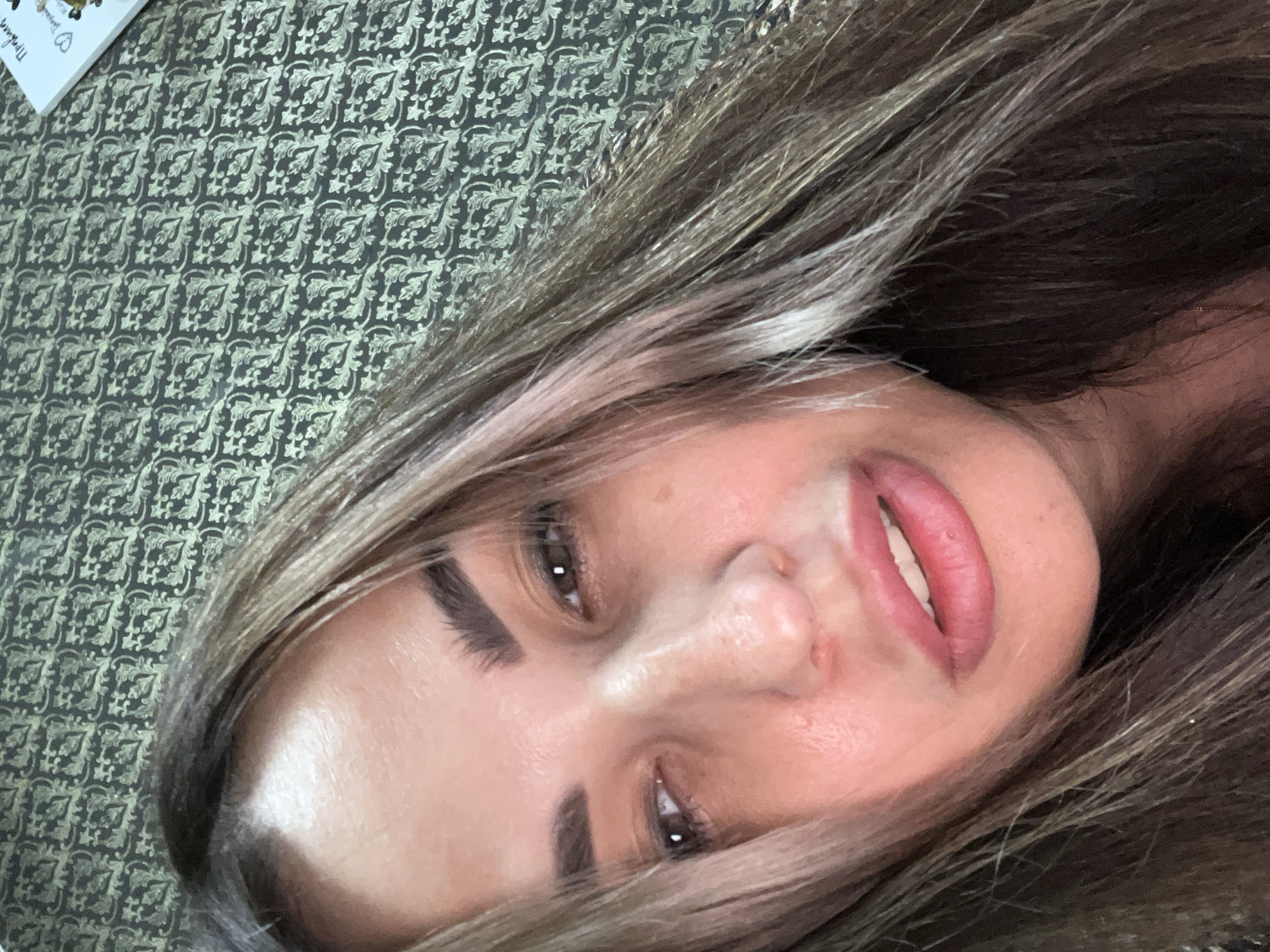 A close-up, head-and-shoulders selfie of a woman with long, dark brown and blonde-streaked hair is taken from a low angle. She is positioned in the center of the frame, her face slightly tilted to her left. Her eyes are open, with her right eye appearing more in focus than her left. She has dark eyebrows and brown irises. Her lips are slightly parted, revealing her teeth, and are painted with a pinkish-red lipstick. Her skin has a warm, rosy tone. The background is a patterned wallpaper with a repeating floral motif in shades of dark green and light gold. In the upper right corner of the image, a white surface is visible with some writing and small drawings on it.
