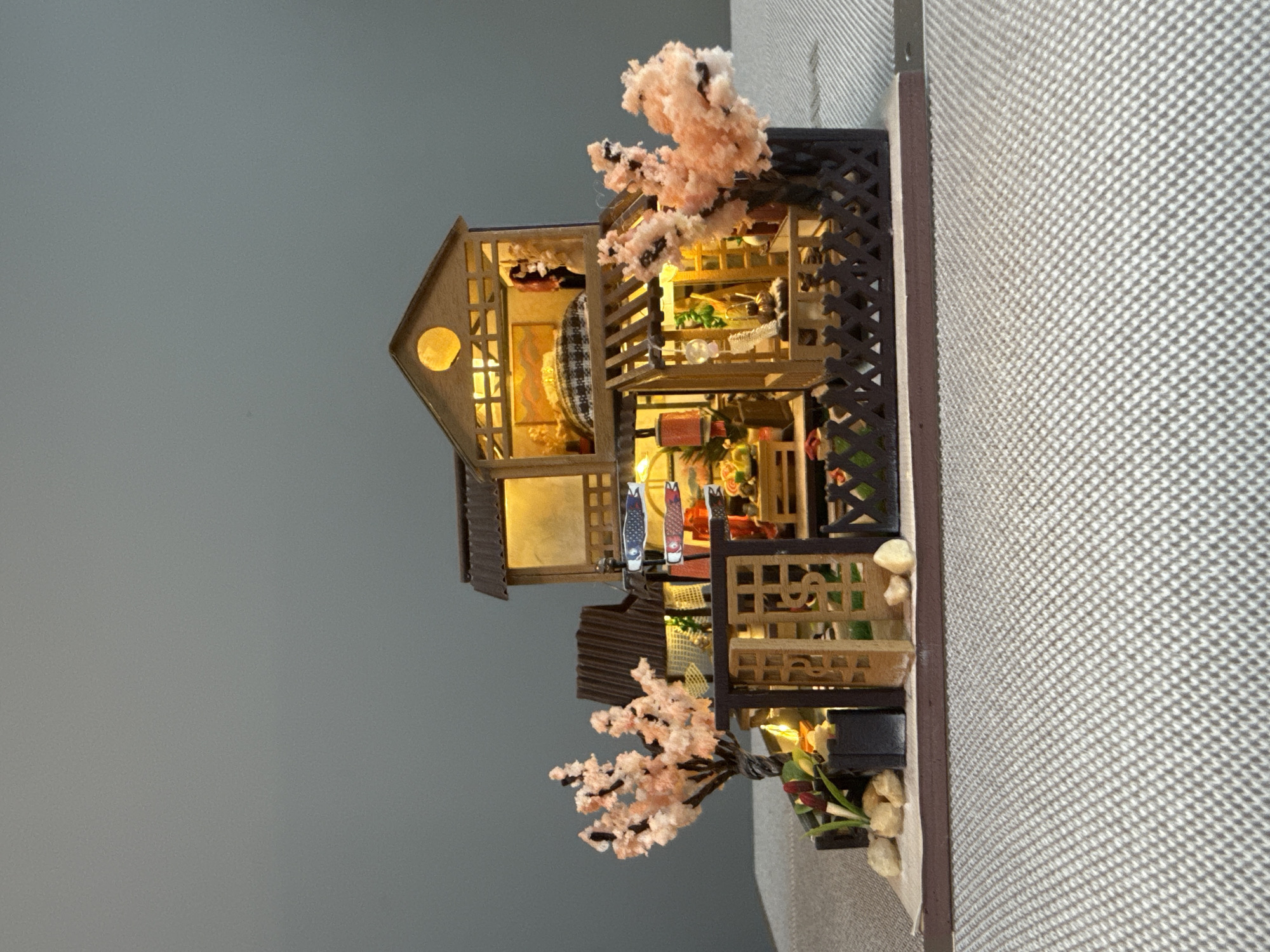The image shows a miniature, two-story building, illuminated from within. The building has dark brown wooden frames and lattice work on the sides. Two flowering trees with pink blossoms are situated on opposite sides of the building. The background consists of a gray wall on the left and a textured white surface on the right.