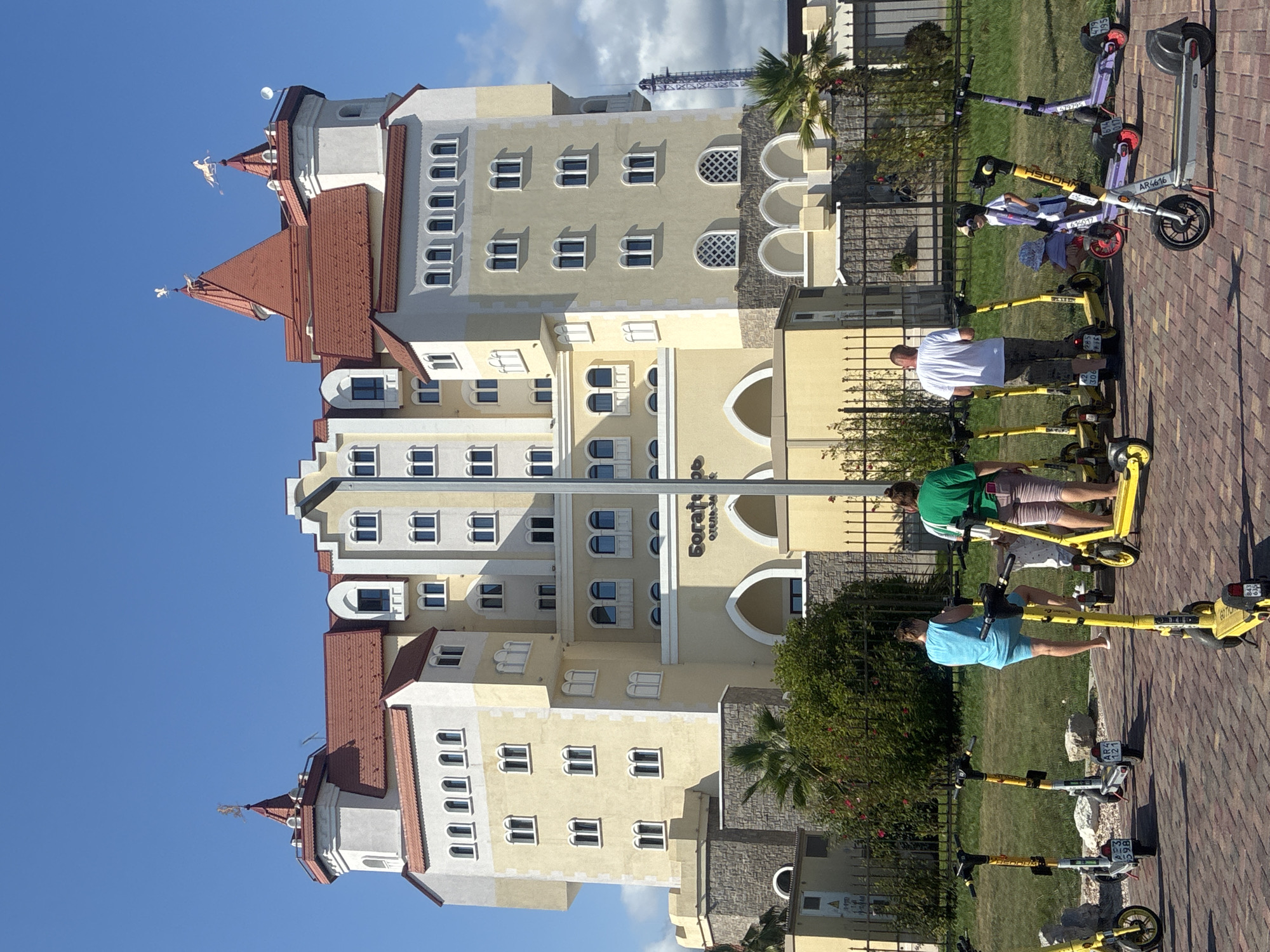 The image shows a large, yellow, castle-like hotel with red roofs against a clear blue sky in Sirius, Russia. In the foreground, several people are standing near a row of parked electric scooters. A woman in a blue top and shorts stands near a yellow scooter, while another woman in a green top and light-colored pants stands next to a yellow scooter, facing away from the camera. A man in a white t-shirt and shorts is also near a yellow scooter. A child in a patterned shirt and a bucket hat is sitting on a small red tricycle, and another child wearing sunglasses and a striped outfit stands beside them. Multiple yellow and a few purple electric scooters are lined up on a brick pathway. A black metal fence encloses a grassy area with bushes and palm trees behind the scooters. The hotel facade features numerous windows and arched entrances, with decorative weathervanes on the towers.