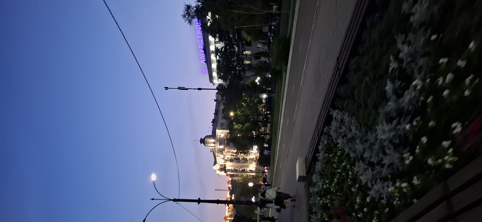 The image depicts a street scene in Blagoveshchensk, Russia, at dusk. A street lamp with a curved arm is visible on the left, and a cell tower stands on the right. Buildings with illuminated windows line the street, and a prominent sign with the word "АМУРСТРОЙ" in blue neon lights is displayed on one of the buildings. Two individuals are walking on the sidewalk in the foreground, facing away from the camera. The street is empty of vehicles. A flowerbed with gray and white foliage and small white flowers is in the lower part of the image, bordered by a dark bench. The sky is a deep blue, transitioning to a lighter hue near the horizon.