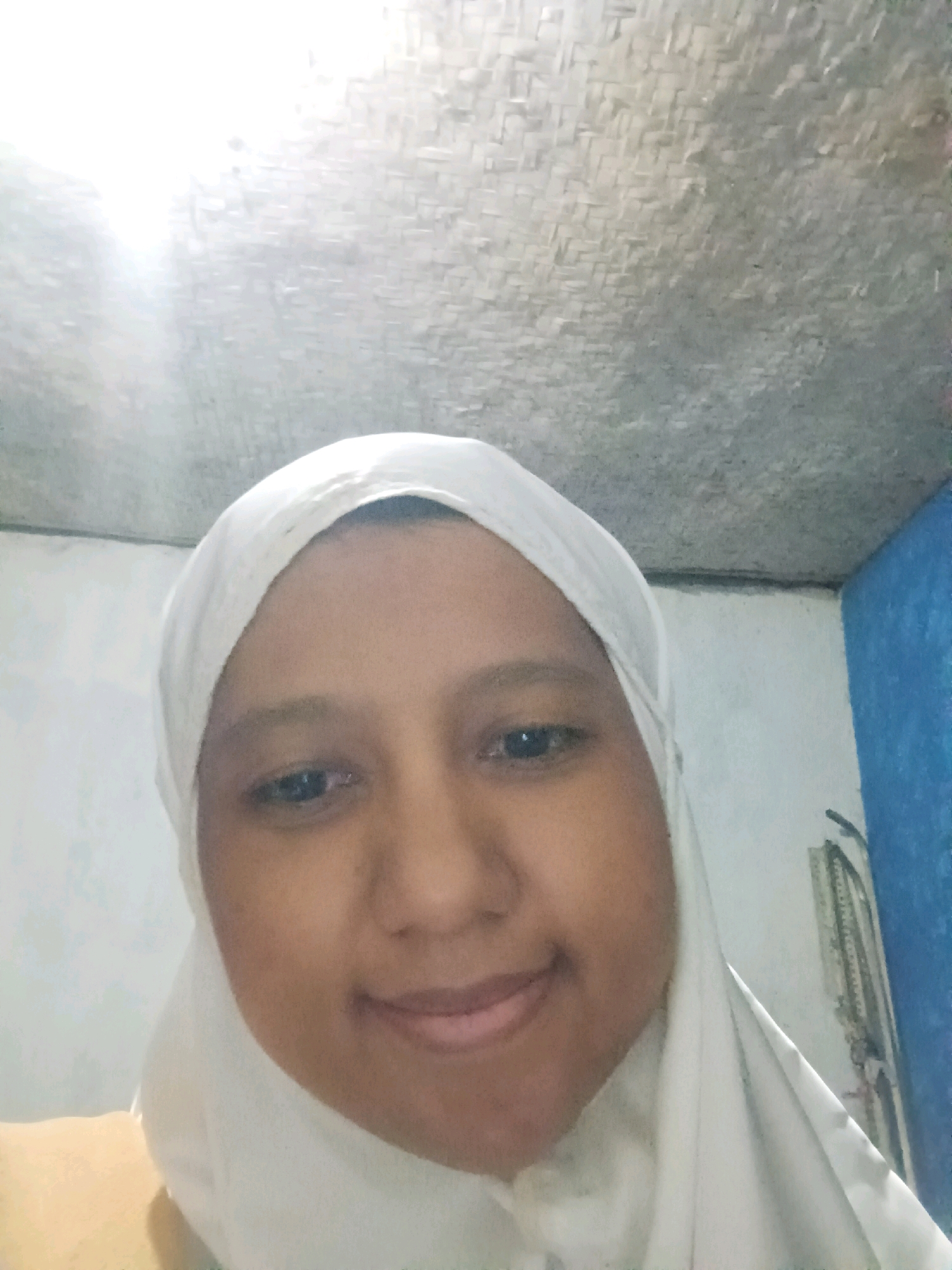 A selfie is taken from a low angle of a young person wearing a white hijab. The person is smiling and looking at the camera. The background appears to be indoors, with a textured ceiling above and a wall to the right. A bright light source is visible at the top left of the image. The overall impression is casual and personal.