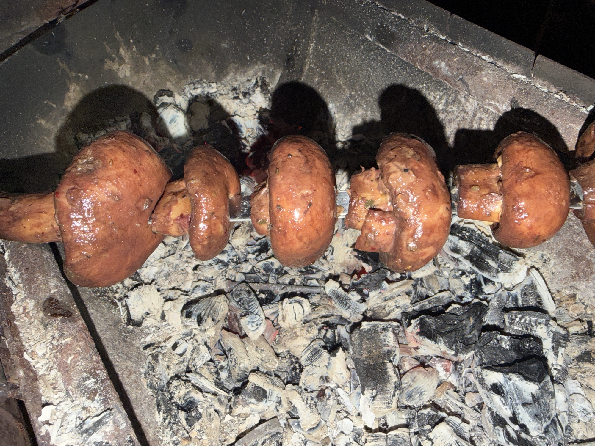 A skewer of five whole, raw mushrooms is positioned vertically above a bed of hot coals and ash in a rustic grill. The mushrooms are a rich brown color and appear to be coated in a marinade or oil, reflecting light from the fire. They are threaded onto a metal skewer that passes through their stems. The coals are white and gray with patches of black char, indicating recent or ongoing use. The grill itself is made of dark, textured material. The background is dark, suggesting an indoor or nighttime setting. There are no people or other living beings visible in the image.
