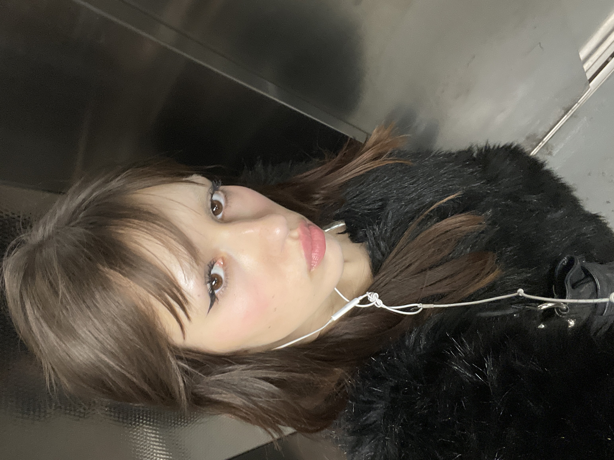 A young woman with brown hair and fair skin poses in an elevator, likely in London, United Kingdom, given the urban backdrop implied by the elevator. She is wearing a black fur coat and has wired earphones connected. Her face is tilted towards the camera, showing makeup with a defined eyeliner and pink lipstick. The elevator has a modern, metallic interior.