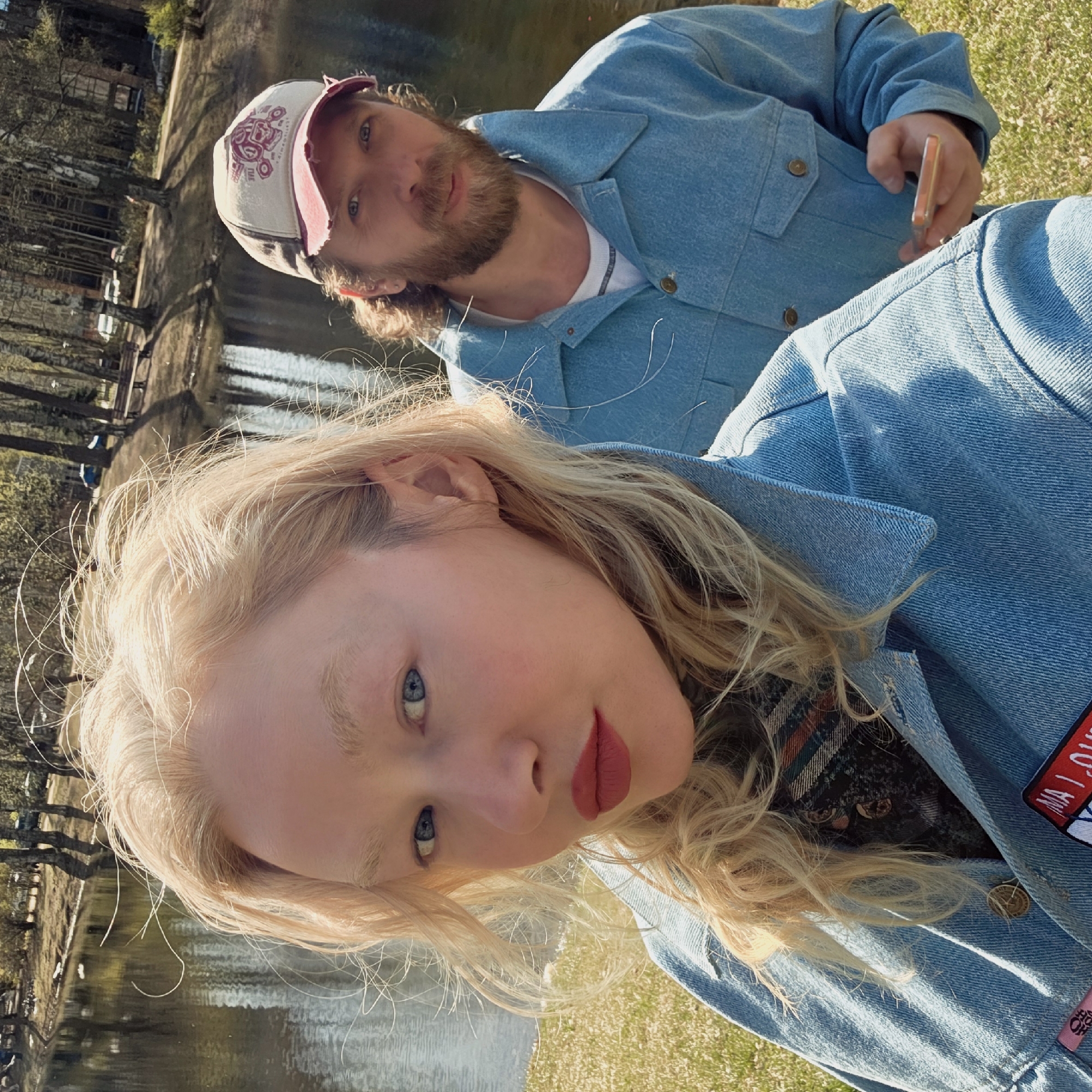 An outdoor, slightly tilted portrait-style photograph captures two individuals, a woman and a man, by a body of water in what appears to be a park setting in Saint Petersburg, Russia, during bright daylight.
In the foreground, a woman with long blonde, wavy hair, fair skin, and blue eyes looks directly at the camera. She has rosy cheeks and is wearing red lipstick. She is dressed in a light blue denim jacket over a dark plaid garment, visible at her collar and side. A red, white, and blue rectangular patch, partially obscured, is visible on her right shoulder of the denim jacket, possibly displaying text like "MA LOV" or "MALO."
Behind and slightly to her right, a man with a beard, light brown hair, and blue eyes also looks towards the camera. He wears a white baseball cap with a graphic design and a light blue denim jacket over a white t-shirt. He is holding a phone with an orange-tinted case in his right hand.
The background features a tranquil body of water, likely a lake or river, reflecting the sky and surrounding elements. Green grass covers the immediate foreground and parts of the bank. Trees, some appearing bare or with early spring foliage, line the far side of the water, where several park benches are also visible. The overall mood is casual and relaxed.