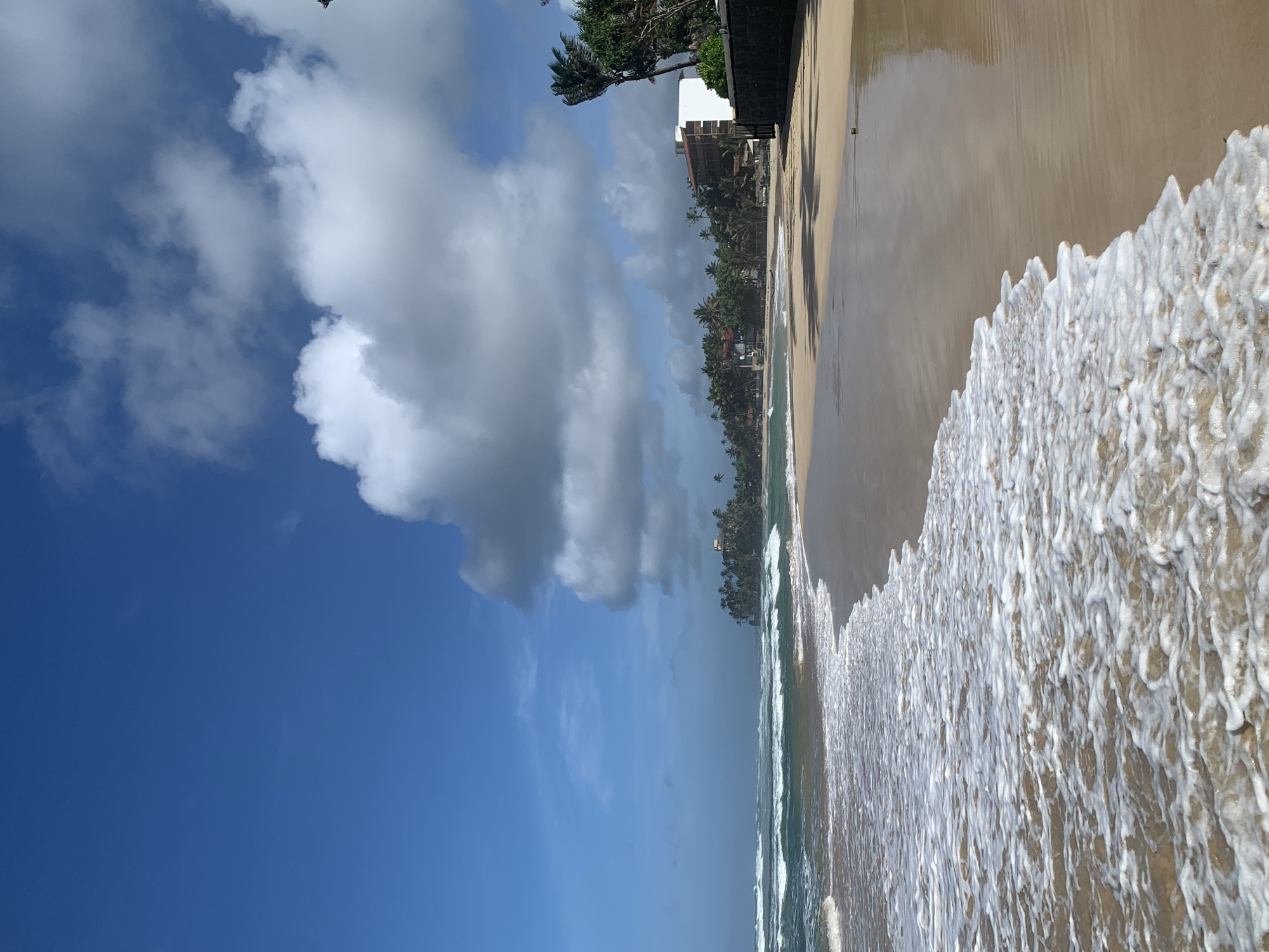 A tropical beach scene unfolds under a partly cloudy blue sky. The foreground is dominated by the ocean's edge, where foamy white waves gently recede from the tan sand. The wet sand reflects the sky and clouds. In the mid-ground, the beach stretches out, lined with lush green palm trees and tropical vegetation. Several buildings, appearing to be hotels or resorts, are nestled amongst the trees. The buildings are modern in design, with some featuring balconies. A dark stone wall runs along the edge of the property closest to the beach. The ocean in the background features rolling waves. There are no people visible in the image.