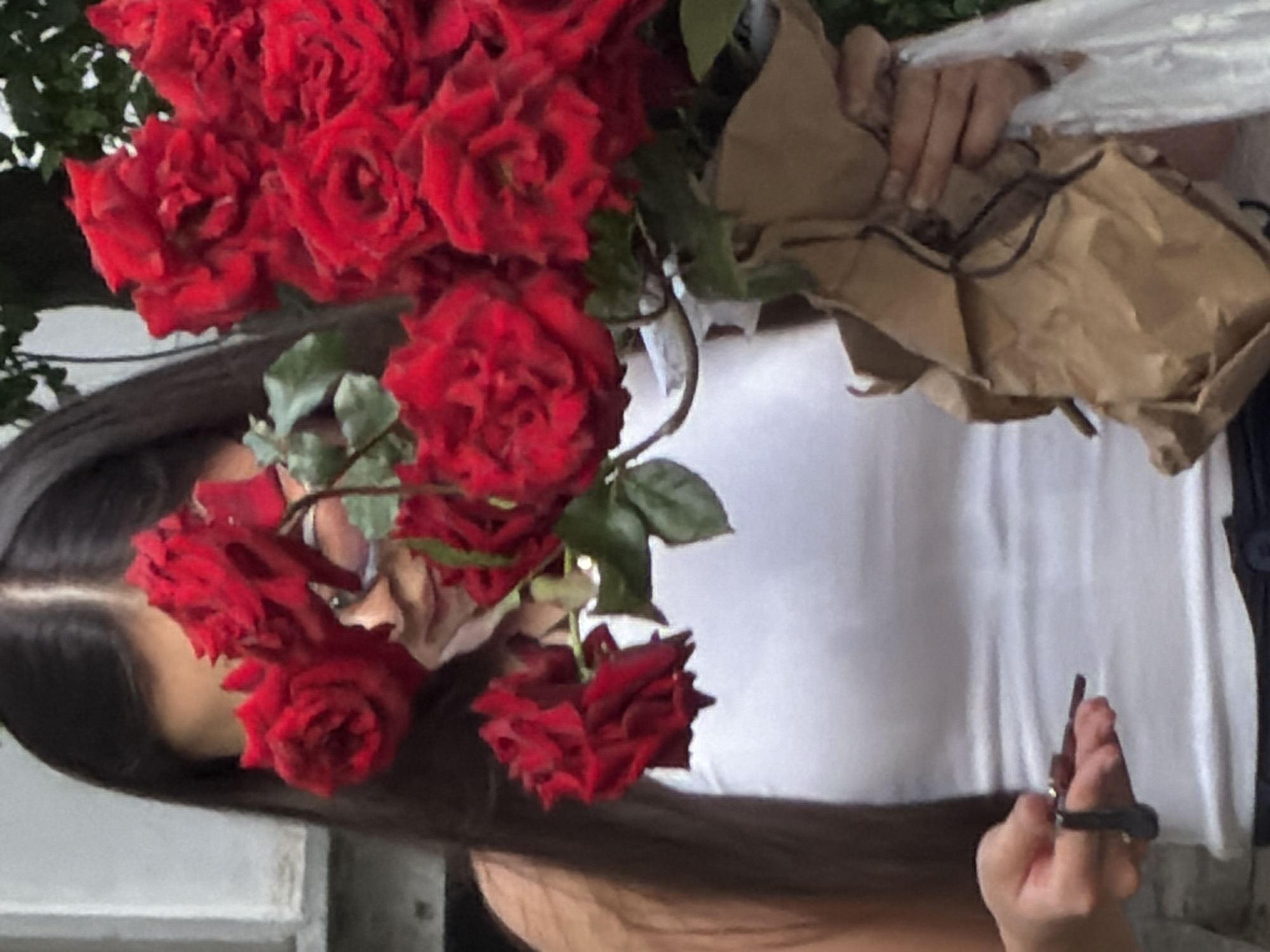 A young woman with long dark hair and glasses, wearing a white tank top, stands in an outdoor setting holding a large bouquet of red roses wrapped in brown paper. Her left hand is visible, holding a small object that appears to be a pair of scissors. The background shows a light-colored wall and greenery.